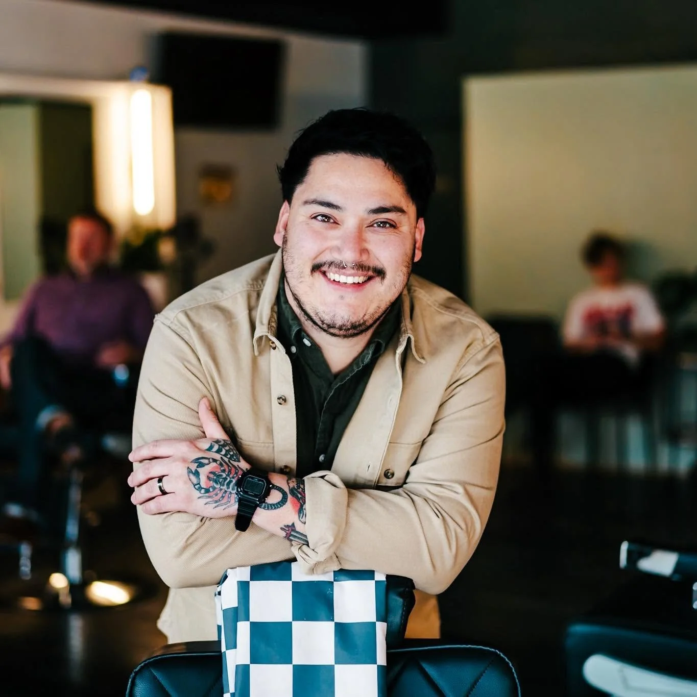 A smiling man with dark hair, tattoos, and a septum piercing, wearing a beige jacket, sitting at a table in a casual indoor setting. There are two blurred people in the background and a checkered bag on the table.