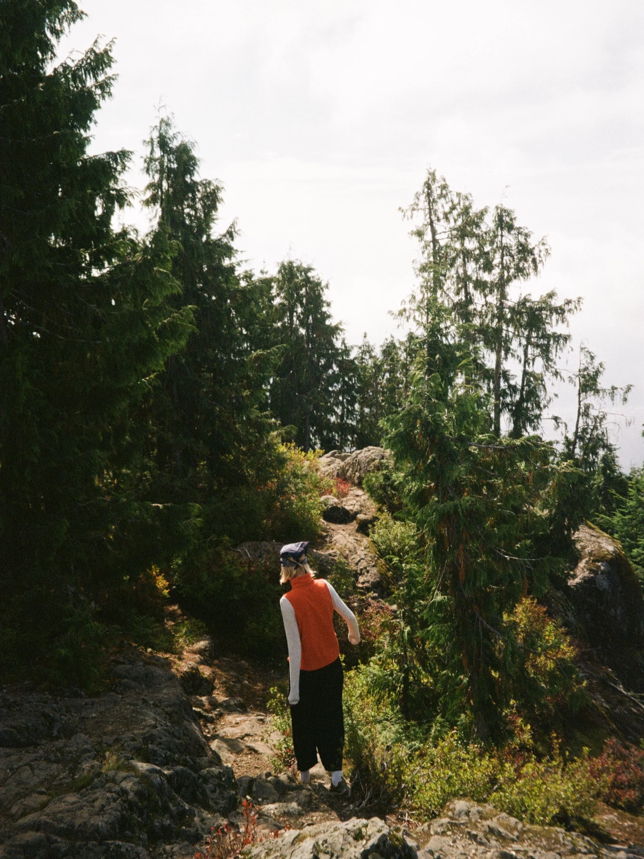 Person in an orange and white jacket hiking on a rocky trail in a forested area with tall trees and greenery.