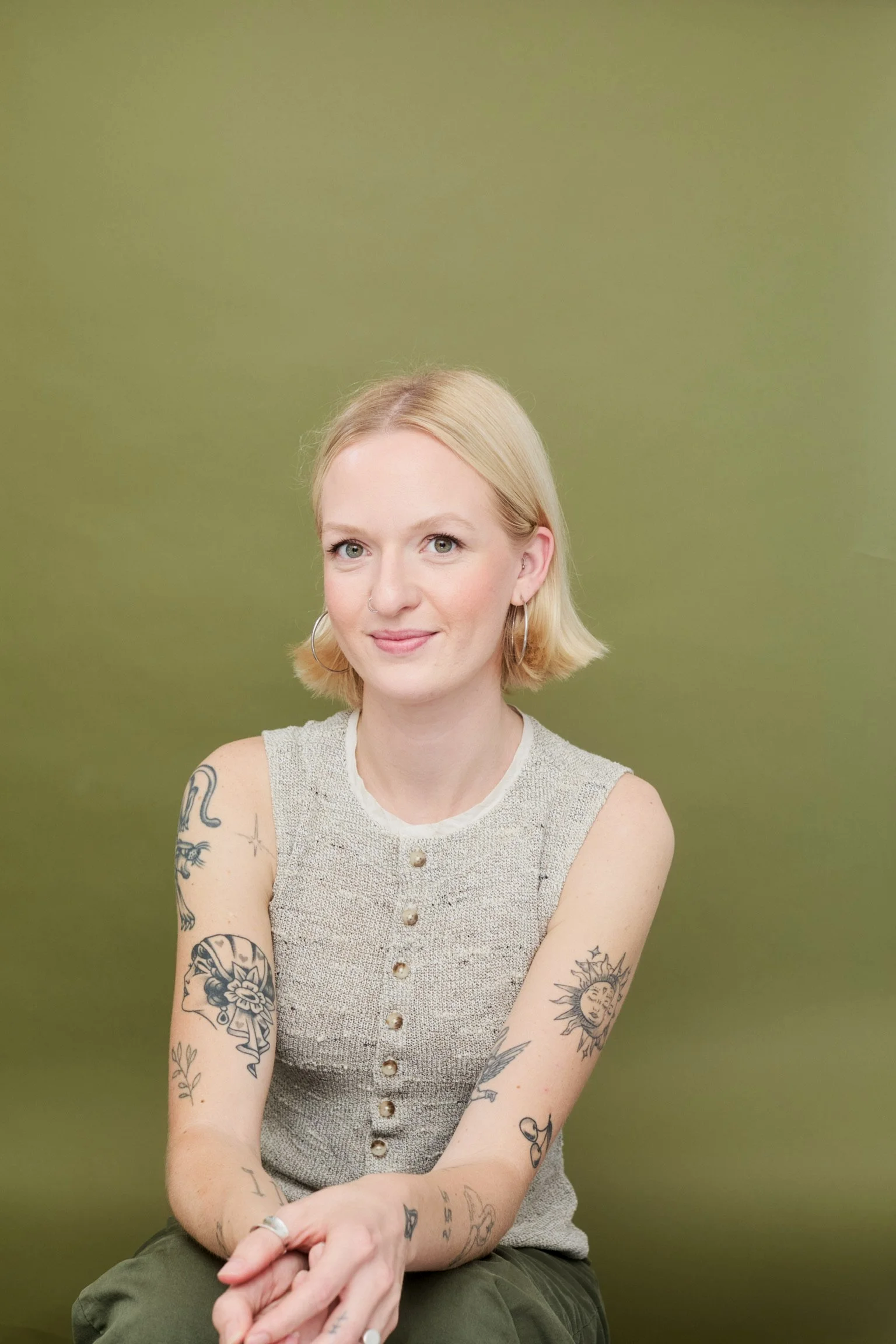 A young woman with short blonde hair, tattoos on her arms, wearing a sleeveless buttoned beige top, sitting against a plain green background.