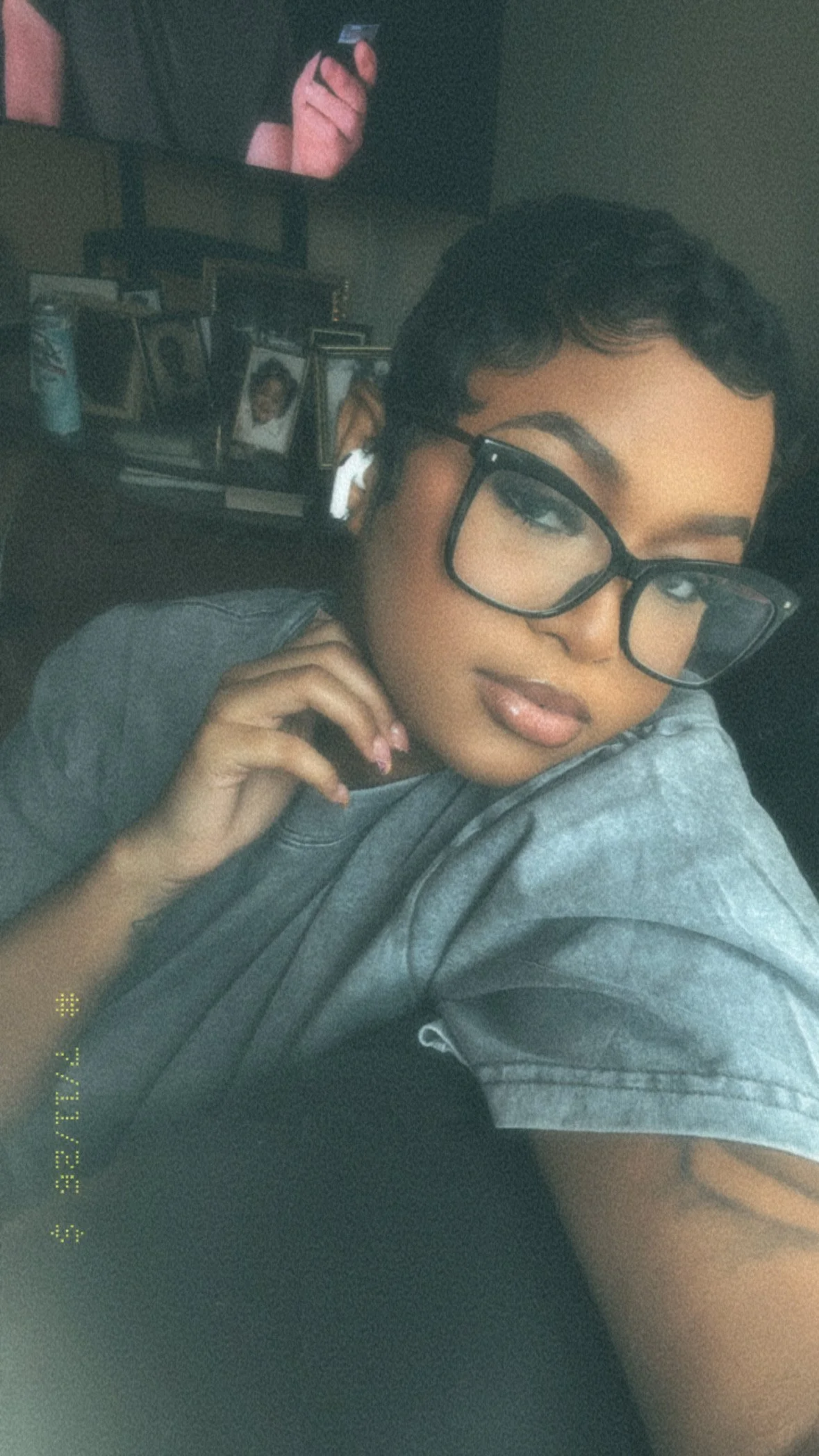 A young woman with short dark hair, wearing large black glasses and a gray shirt, resting her chin on her hand and looking at the camera. In the background, there is a desk with framed photos and a television screen showing a hand holding a remote control.