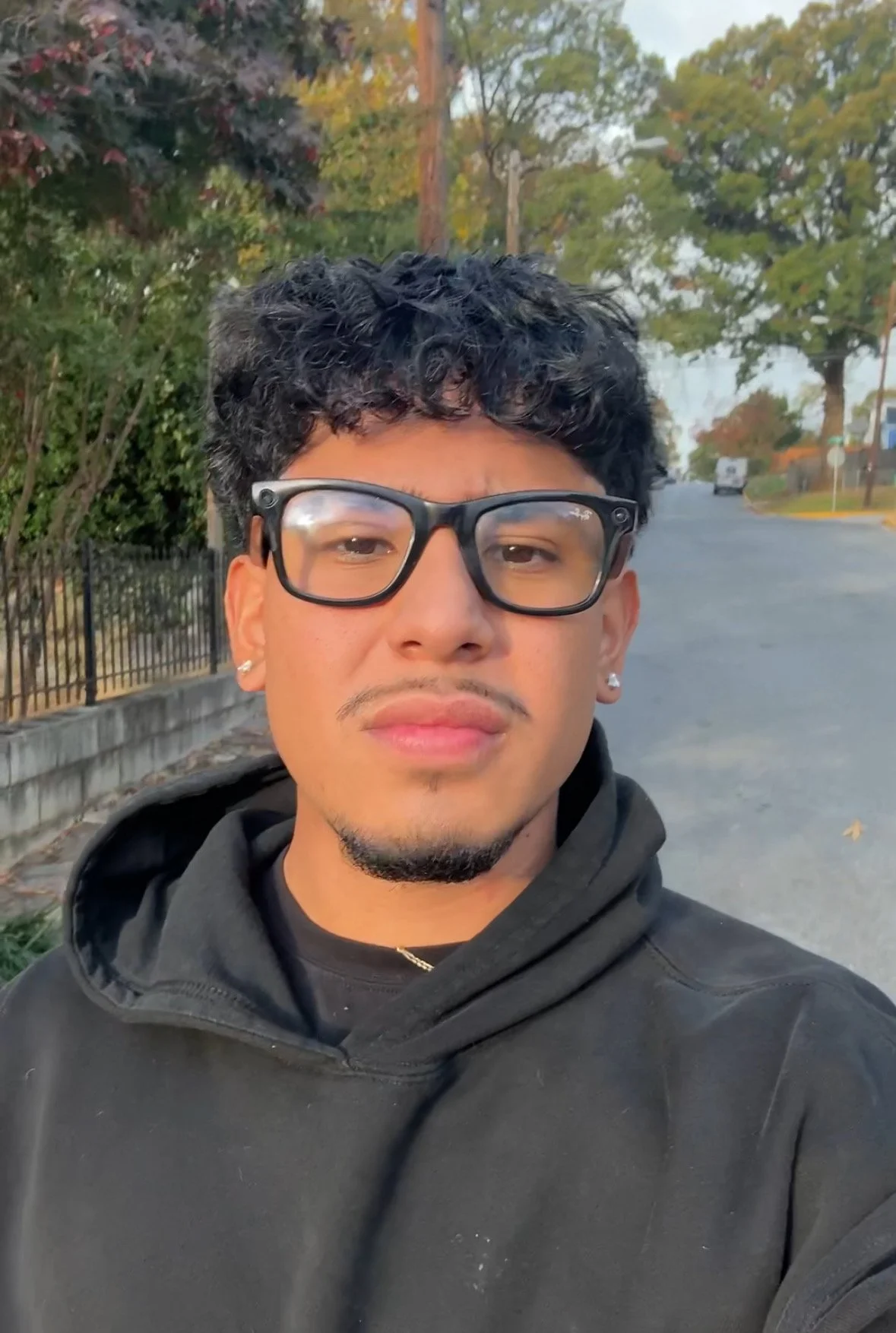 A young man with black curly hair, glasses, earrings, and a goatee taking a selfie outdoors on a cloudy day. He is wearing a black hoodie, and trees with autumn leaves are visible in the background.
