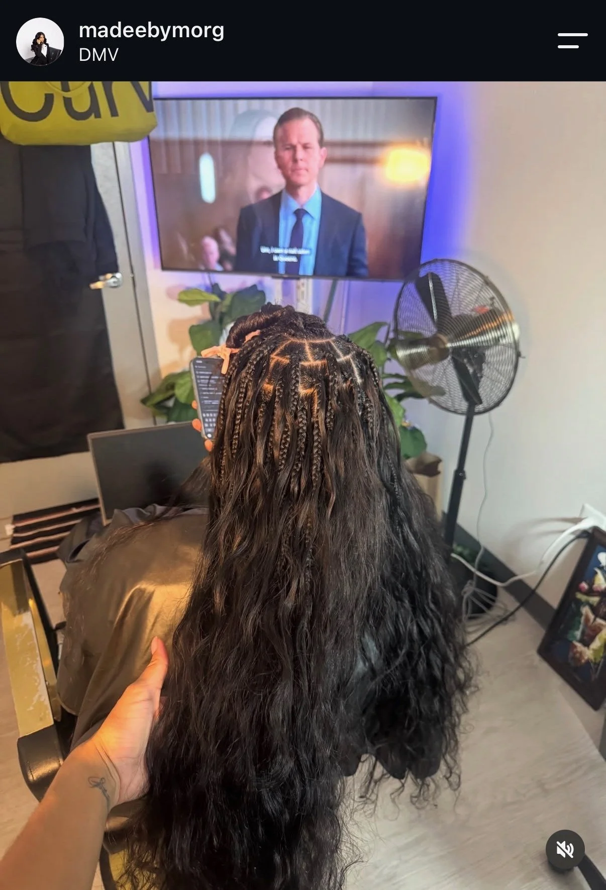 Person with long, curly hair having their hair braided by a stylist while watching a television screen.