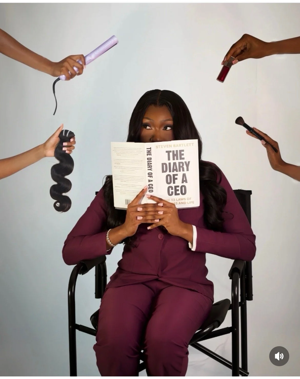A woman in a burgundy suit is sitting in a chair, holding a book titled "The Diary of a CEO." She is surrounded by hands holding beauty tools, including a curling iron, a hairbrush, makeup brushes, and small bottles. The woman is looking slightly to her right while reading the book.