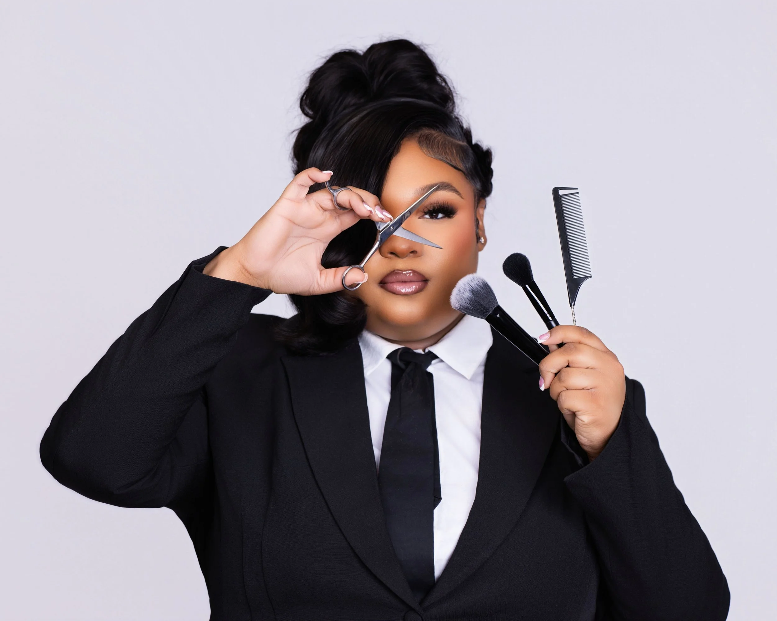 Woman with dark hair styled in an updo, holding scissors and a comb while fixing her hair, dressed in a black blazer and white shirt against a plain light background.