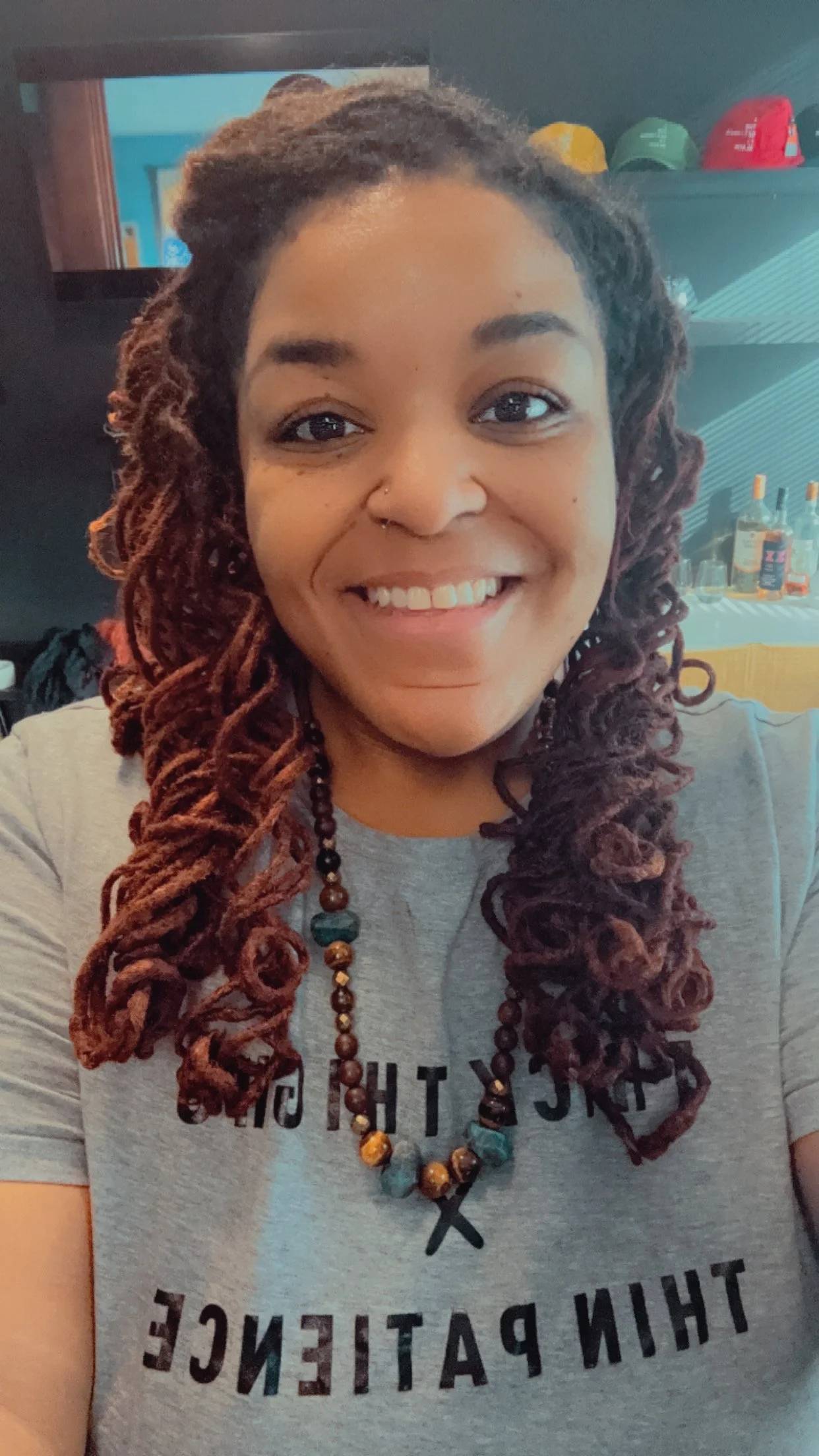 A woman with long, styled dreadlocks and a nose piercing, smiling at the camera. She is wearing a gray shirt with black text and a beaded necklace, standing in a room with shelves of bottles in the background.