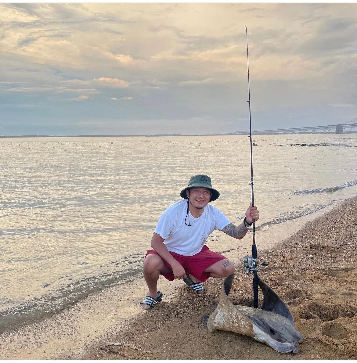 A man kneeling on a sandy beach with a fishing rod, smiling next to a large fish he caught, with water and a bridge in the background.