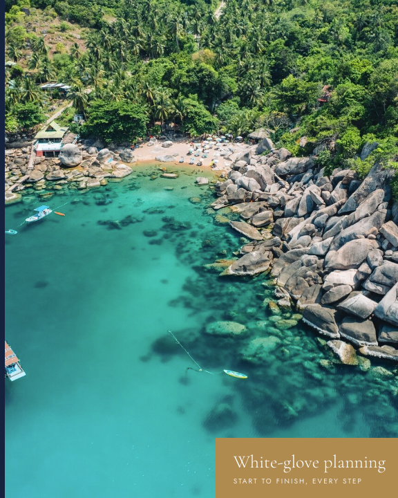 Aerial view of a tropical beach with clear turquoise water, sandy shore with umbrellas, surrounded by lush greenery and large rocks.