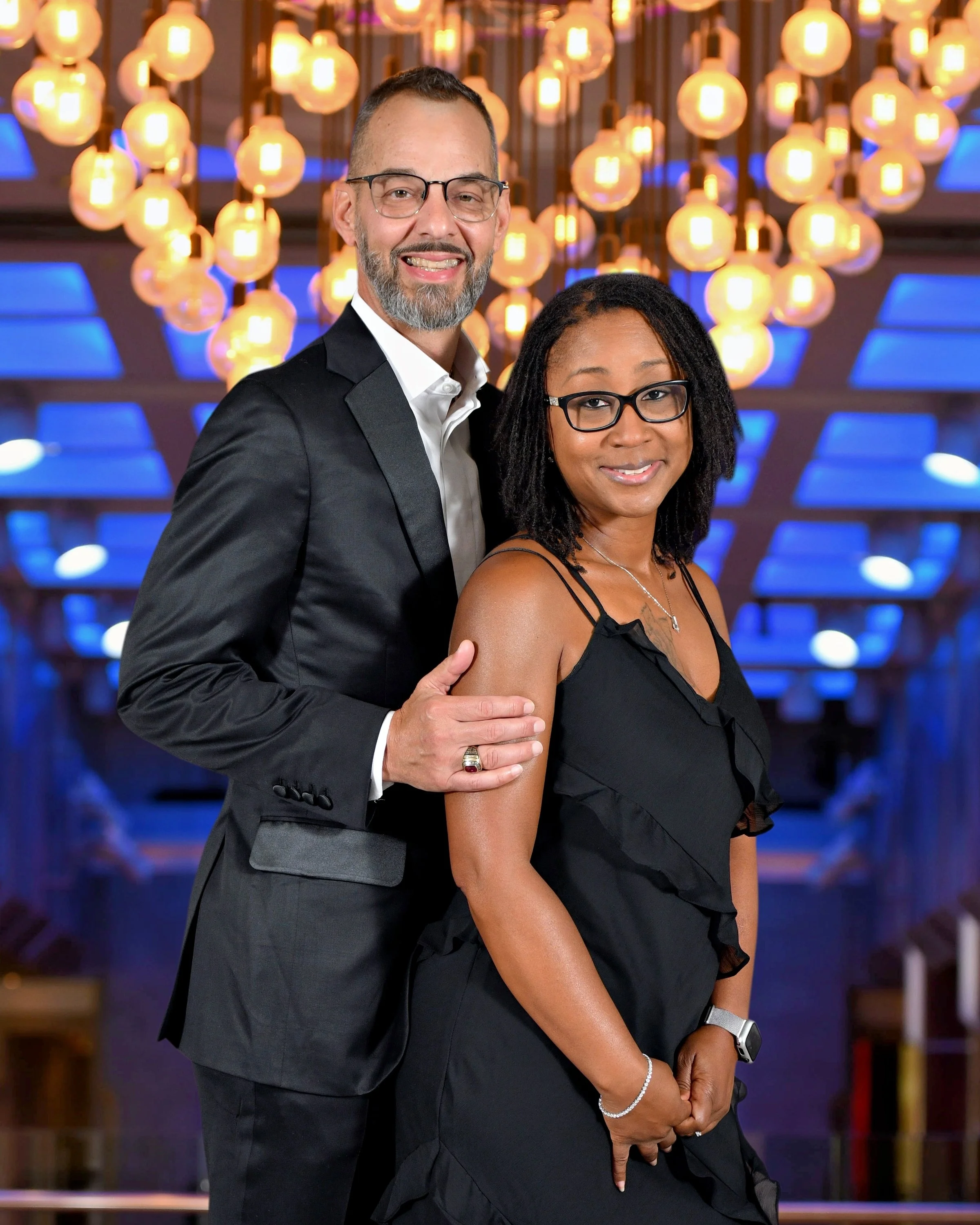 A smiling man with glasses and a beard in a tuxedo and a woman with glasses and a black dress standing close together in front of a decorative background of warm glowing lights and blue accents on formal night on a Royal Caribbean Cruise.