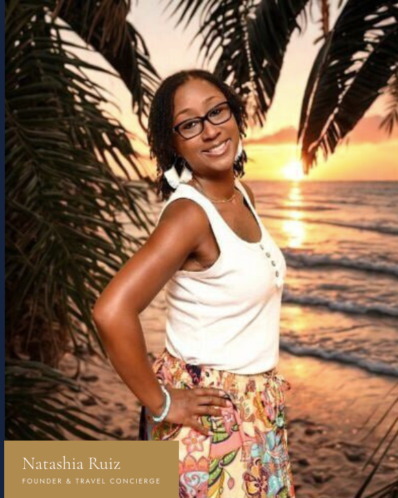 A woman with glasses and curly hair standing on a beach at sunset, surrounded by palm trees, wearing a white sleeveless top and colorful patterned skirt.