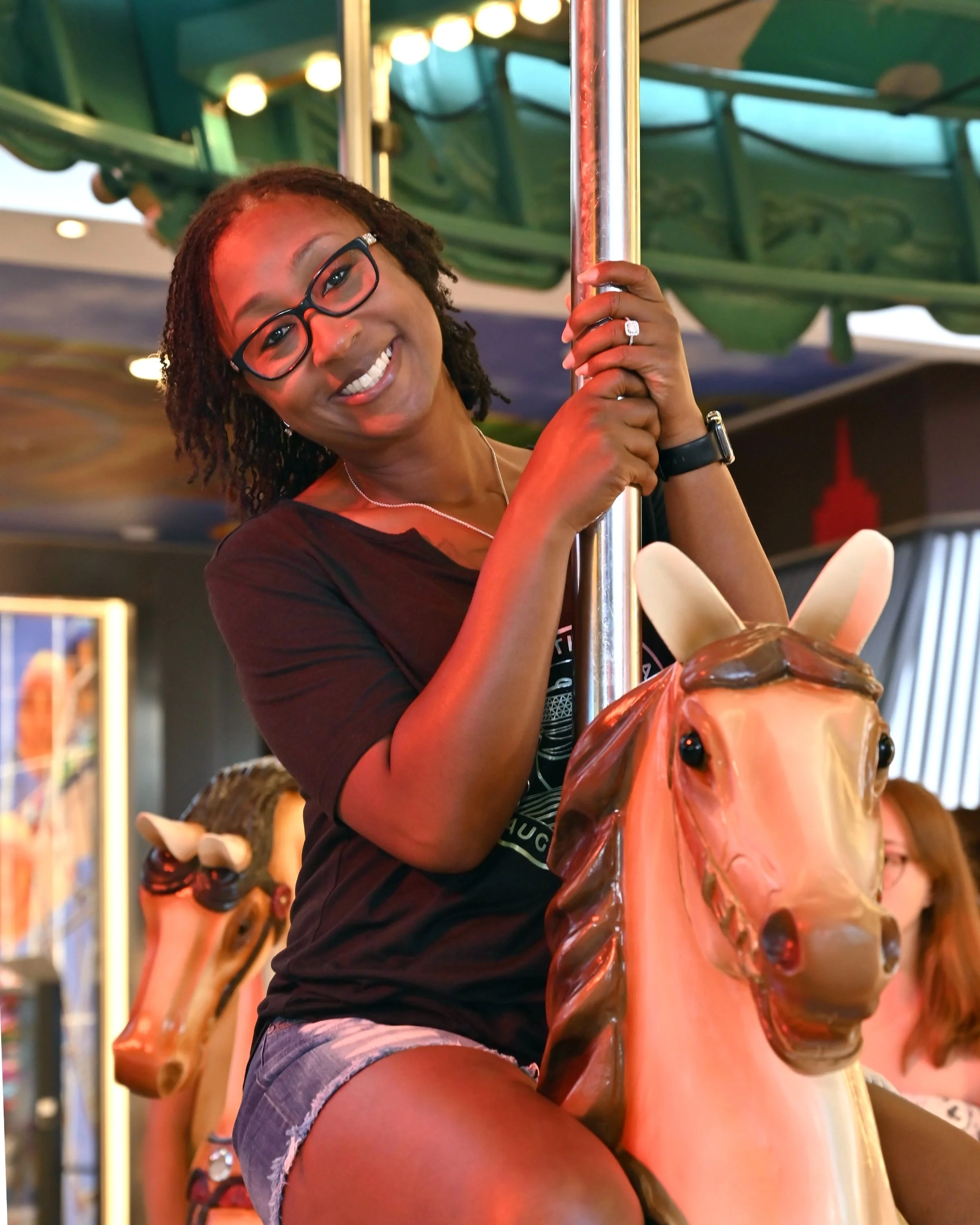 A woman with glasses smiling and holding a pole while riding a carousel horse on a Royal Caribbean cruise.