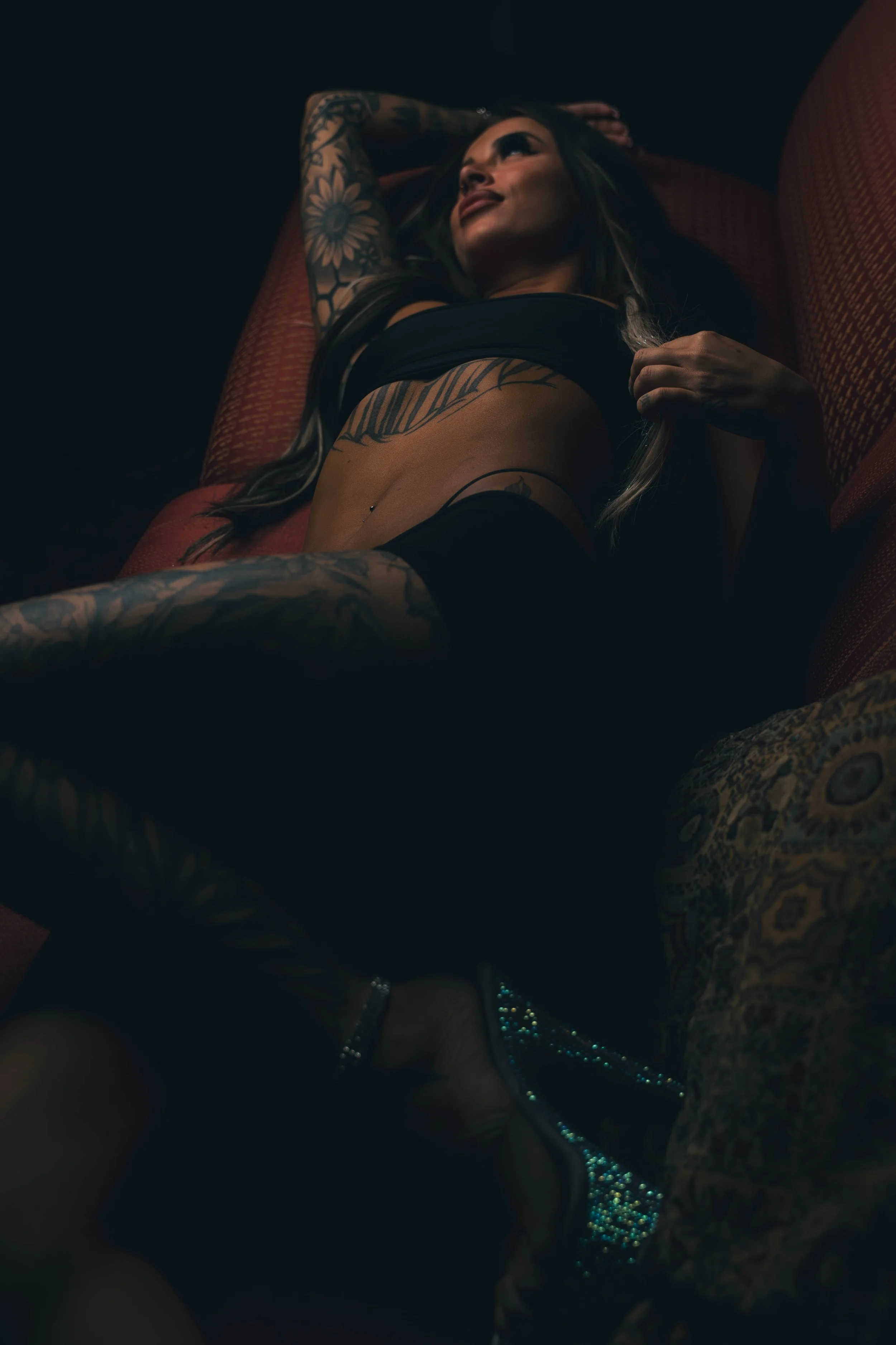 A woman with tattoos on her arm and chest lying on a red couch, looking upward, with dark surroundings.