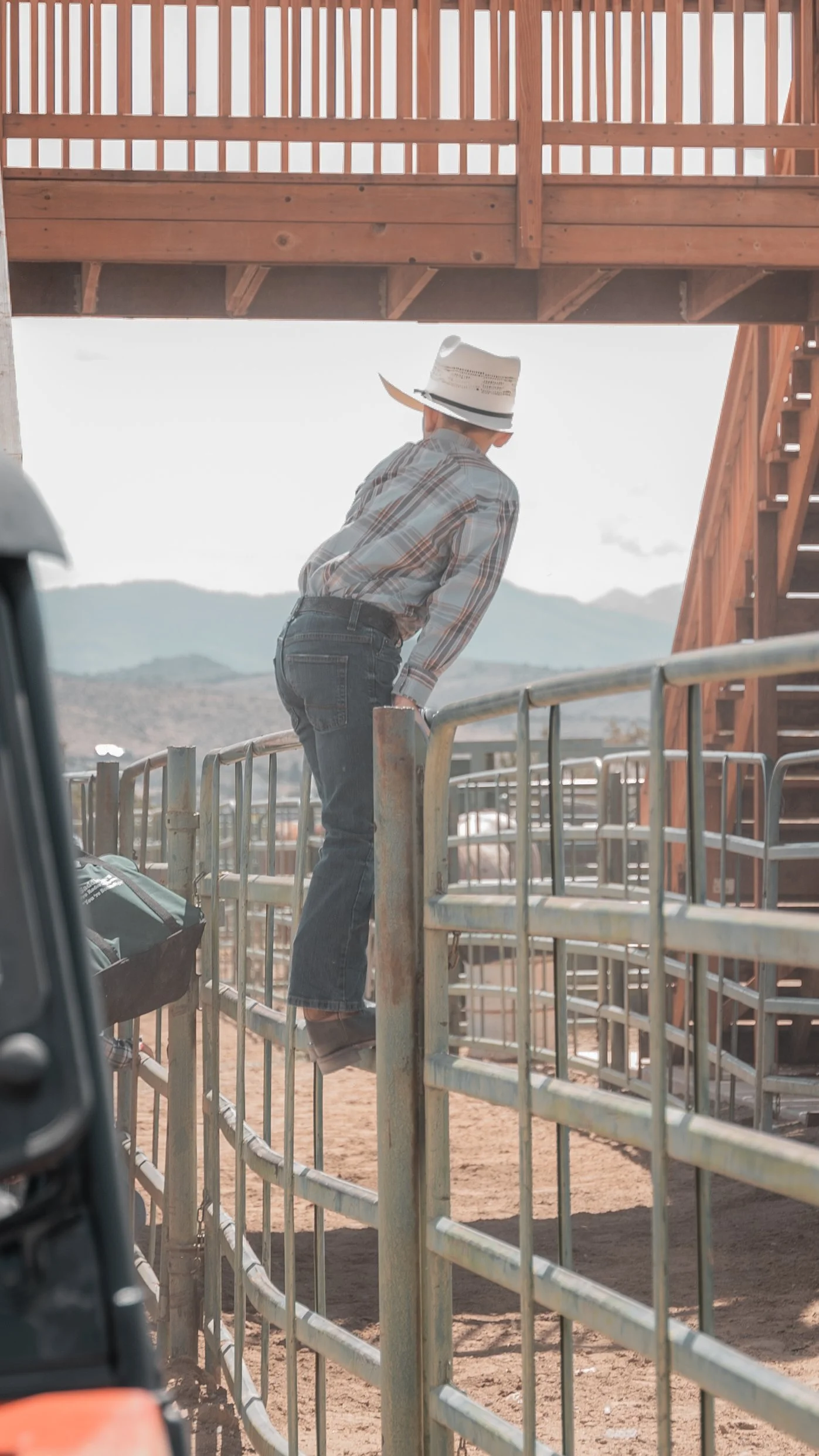 Person wearing a white sun hat, plaid shirt, and jeans standing on a metal railing under a wooden platform, with a desert landscape and mountains in the background.