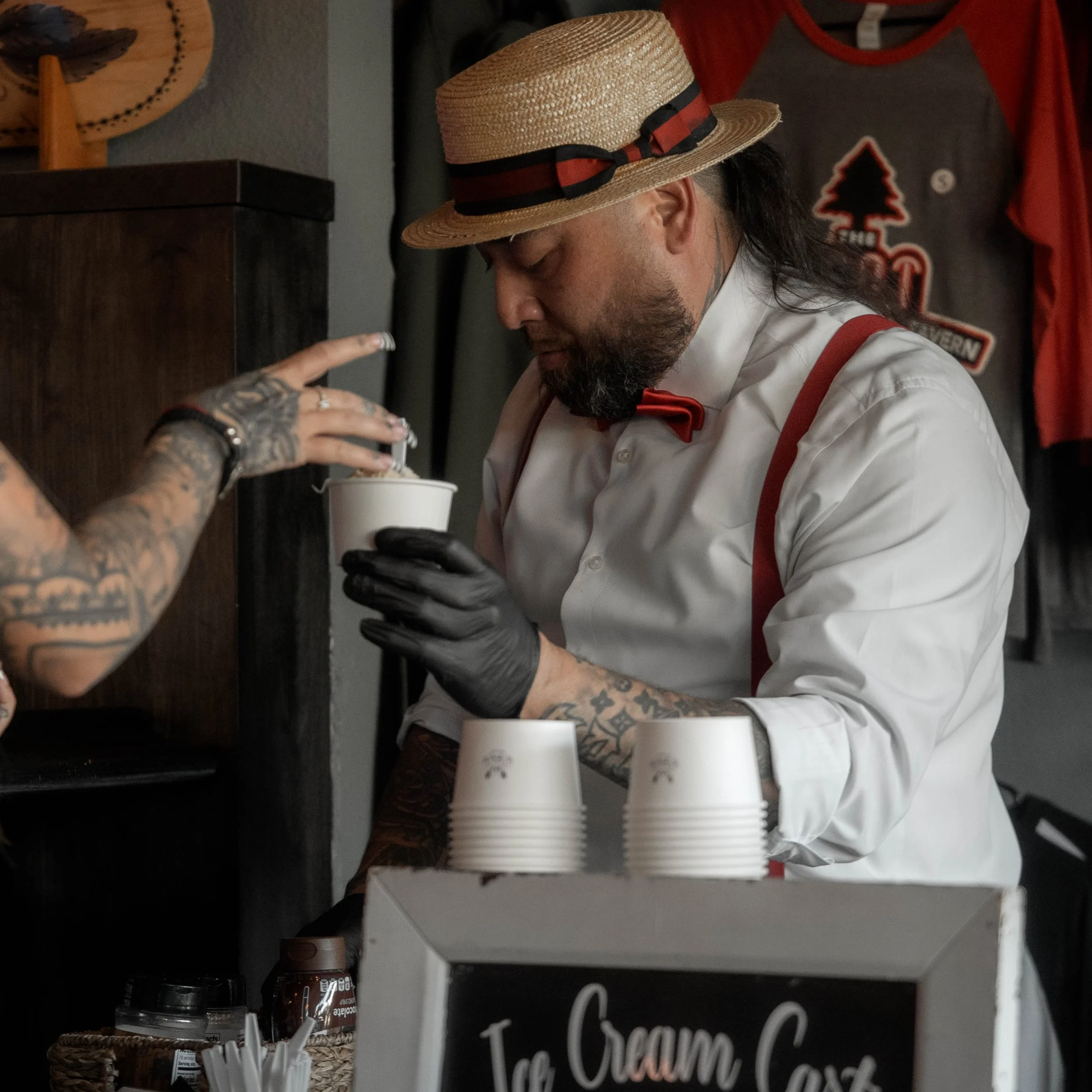 A man with long hair, a beard, and tattoos on his arms, wearing a straw hat with a ribbon and a white shirt with red suspenders, is handling white disposable cups in a setting with a dark wooden counter, a small jar, and a blackboard sign that is par