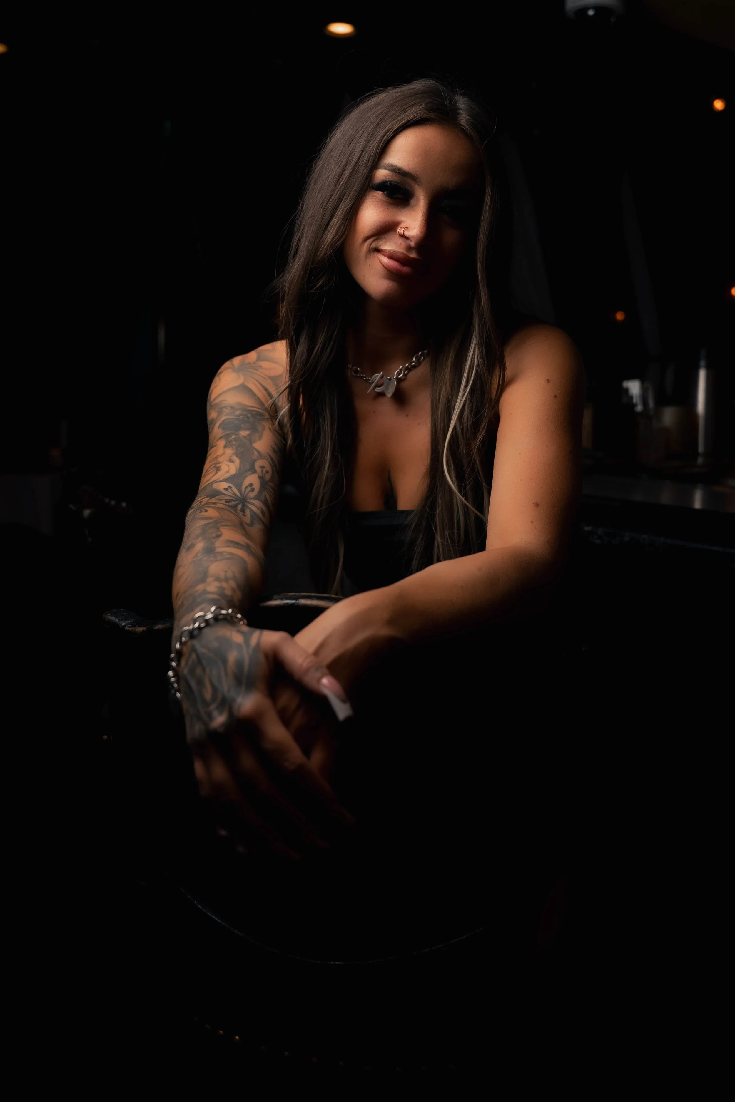 A woman with long dark hair, tattoos on her left arm, wearing a necklace and a black top, sitting at a table in a dimly lit setting with black background and small orange lights.