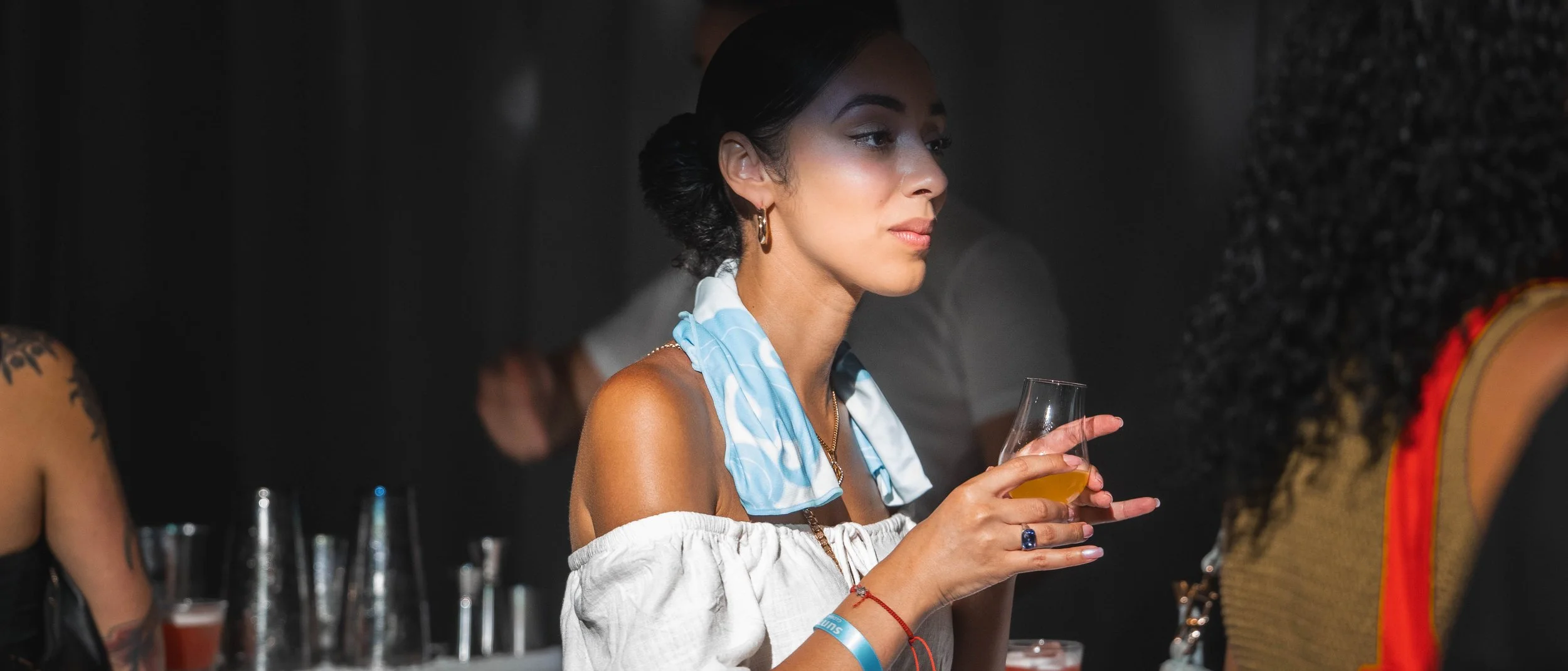 A woman holding a glass of orange-colored beverage in a social setting, wearing a white off-shoulder top and accessories, with dark hair styled in a bun and a blue patterned scarf around her neck.