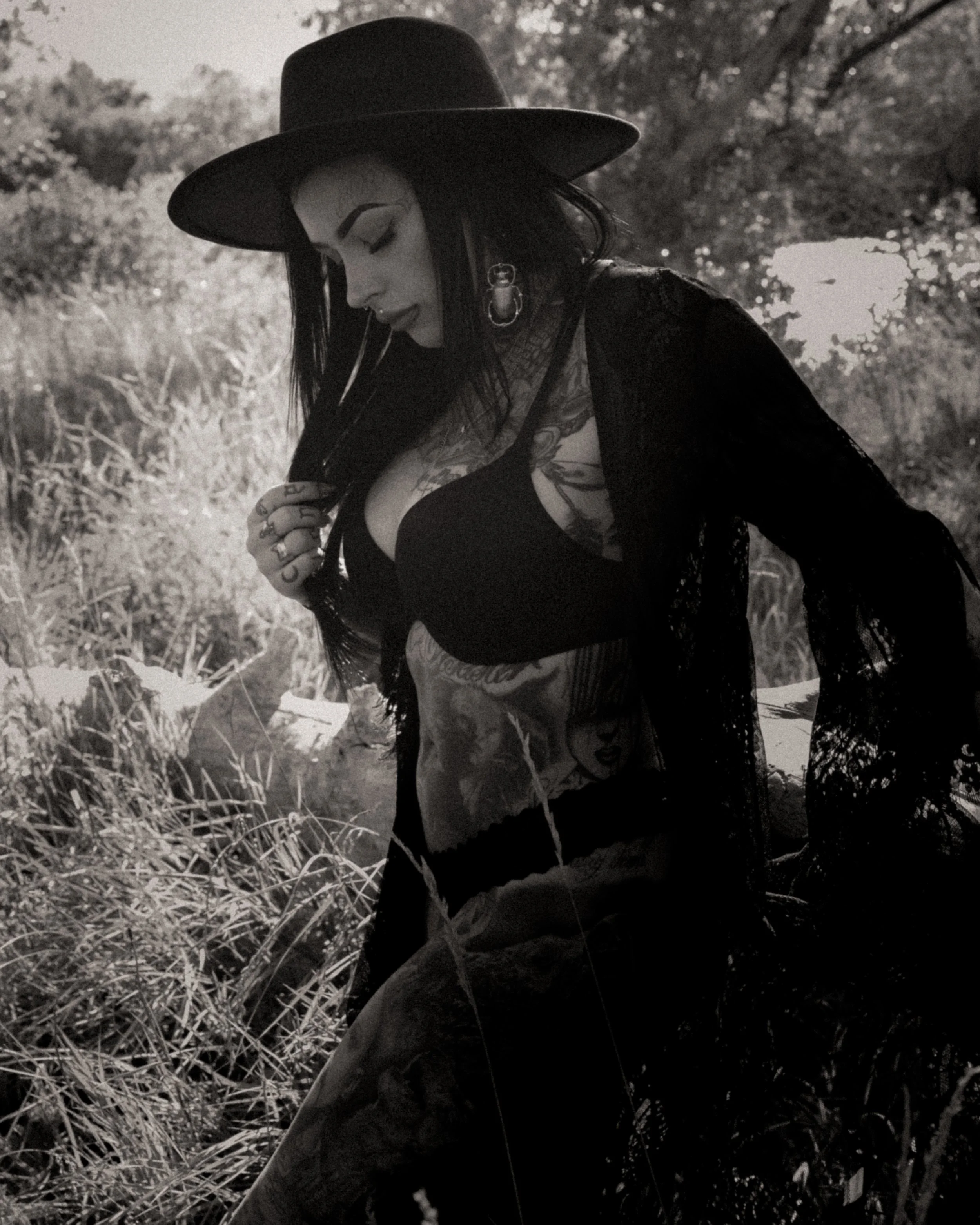 A woman with dark hair and tattoos on her arm, wearing a black top, a ballet-style earring, and a wide-brimmed hat, looking down while standing outdoors in a natural setting with tall grass or plants.