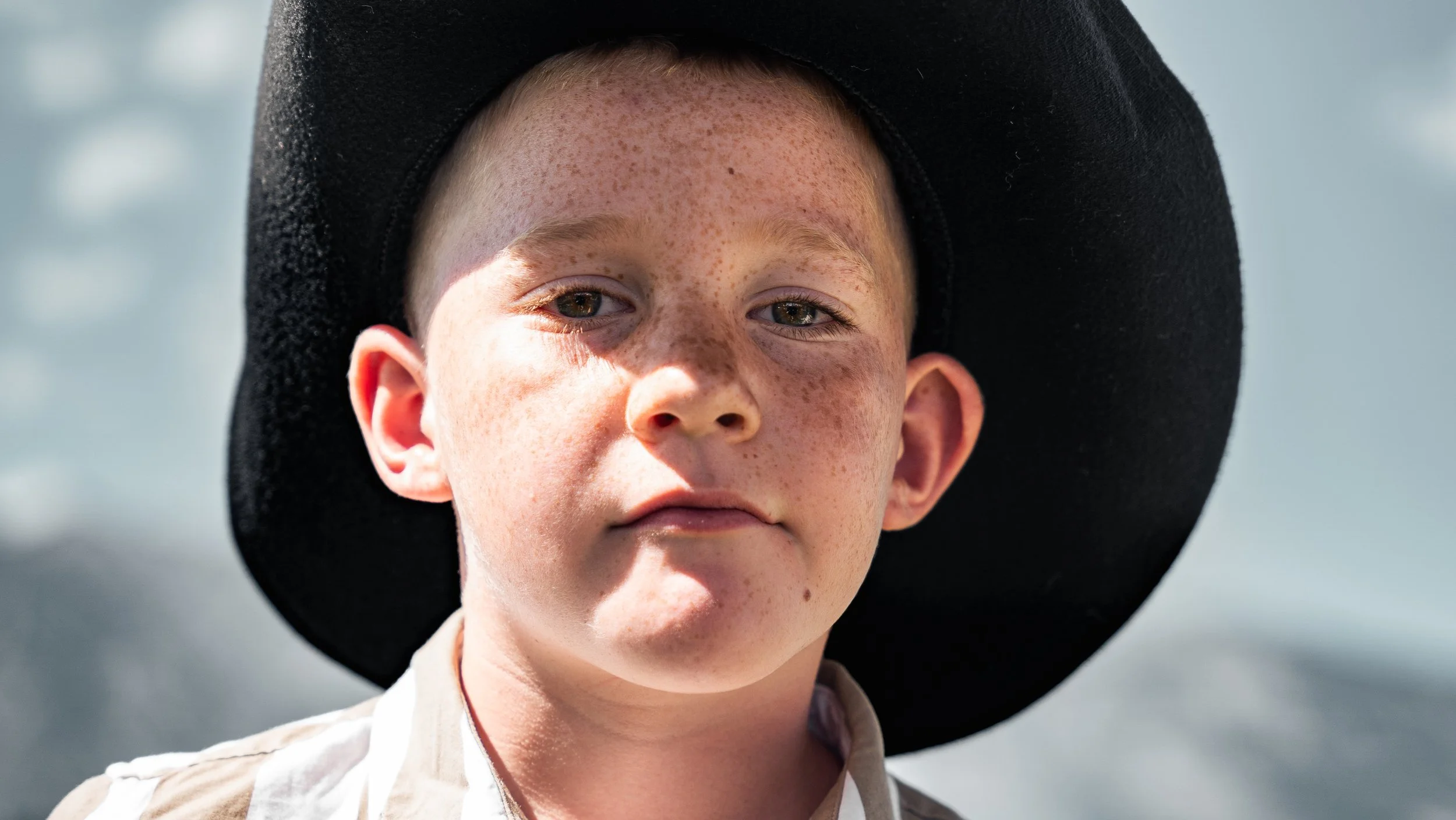 Close-up of a young boy with freckles, wearing a large black hat and a beige shirt, standing outdoors.