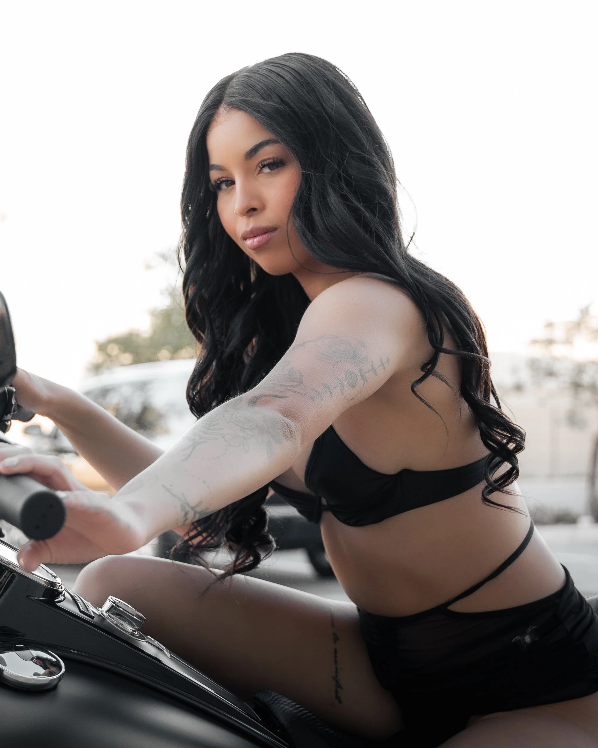 A young woman with long black hair, tattoos, and wearing a black top is riding a motorcycle outdoors. The background is blurred with trees and sunlight.