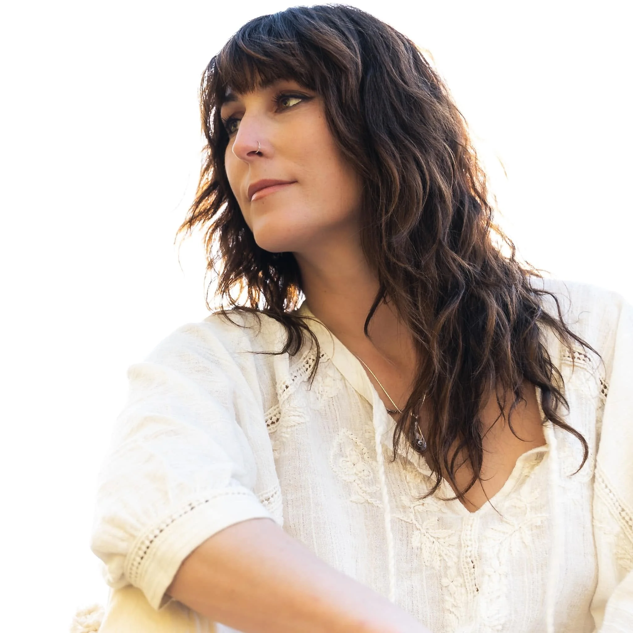 A woman with curly brown hair and a nose piercing, wearing a white crochet top, looking to her left against a bright background.