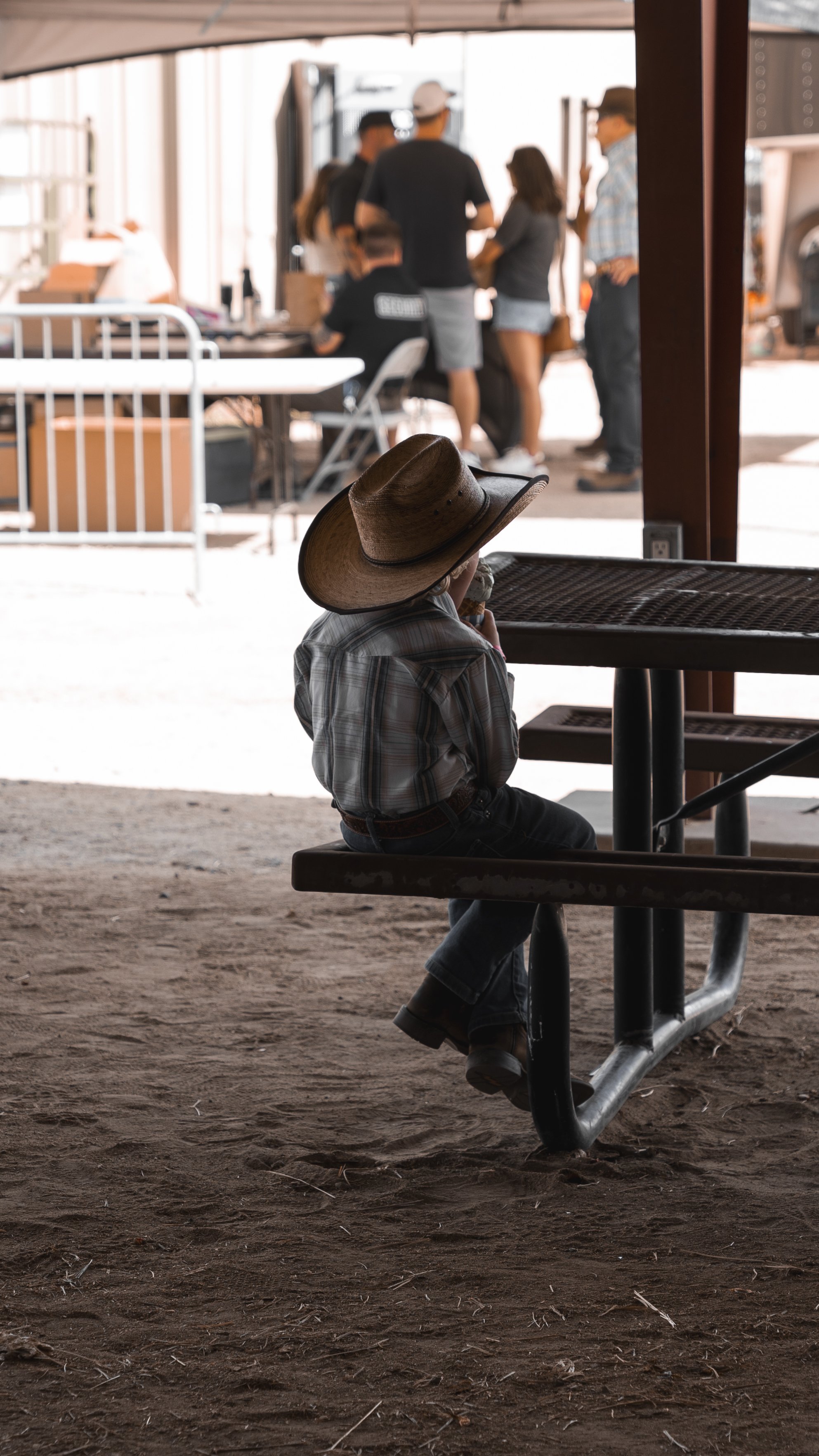 A young boy wearing a plaid shirt, jeans, and a cowboy hat sitting at an outdoor picnic table with his hat pulled low over his face, looking away from a group of people who are gathered around a table in the background.