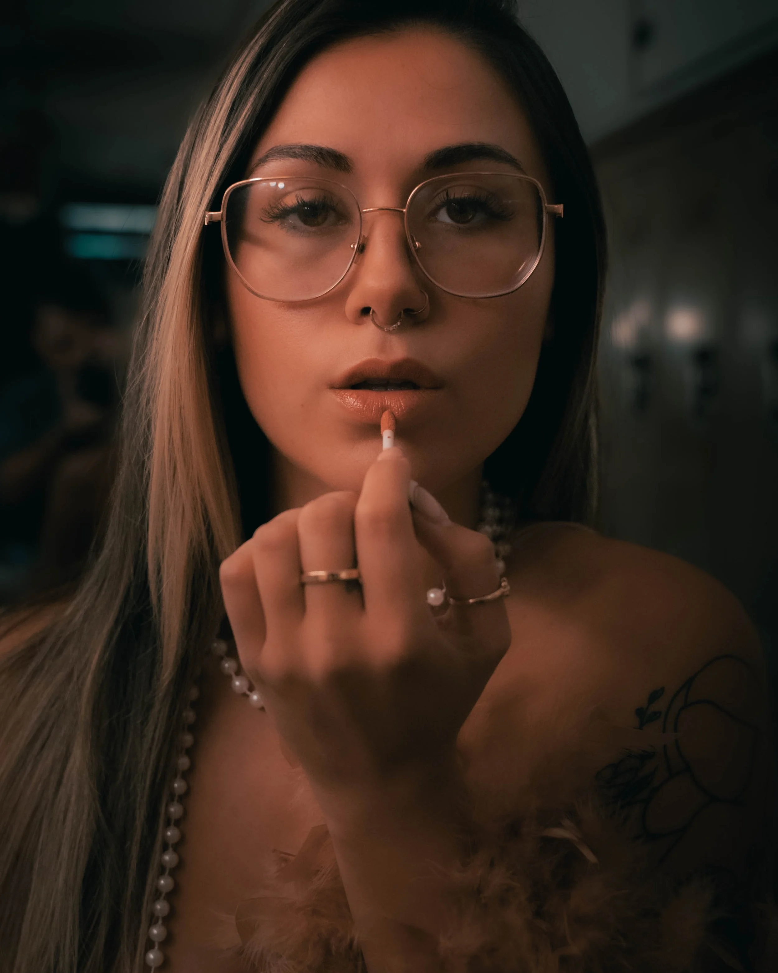 A young woman with long straight hair, wearing large round glasses, a pearl necklace, and a nose ring, is holding a cigarette near her lips. She has a tattoo on her arm and is in a dimly lit indoor setting.