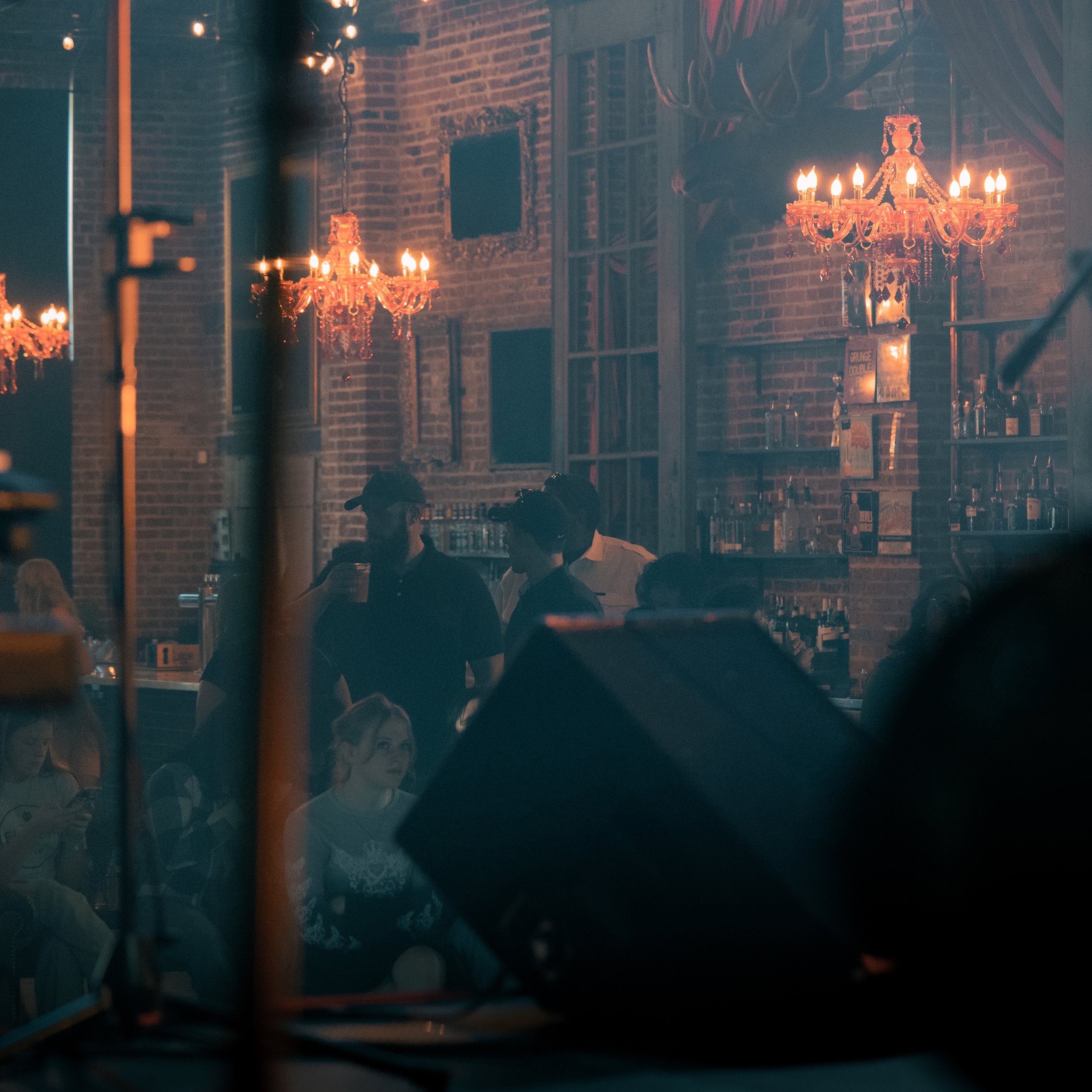 Dimly lit interior of a restaurant with brick walls, ornate chandeliers, and shelves with books. People are seated at tables, some in conversation. The overall ambiance is cozy and vintage.