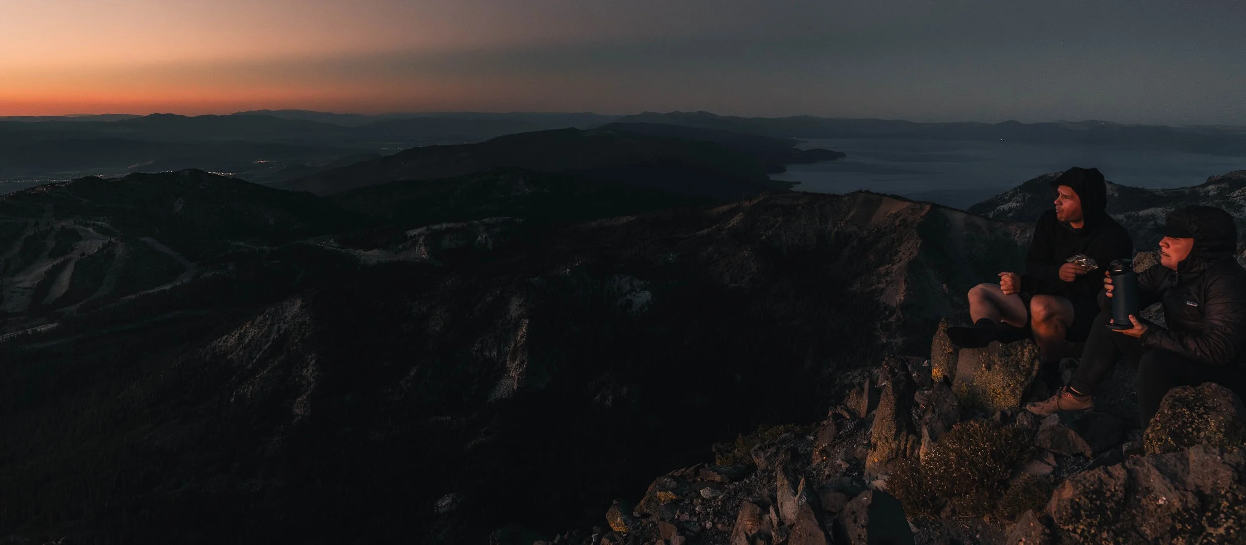 Two people sitting on rocks at the mountain top during sunset, with scenic view of mountains and water in the distance.
