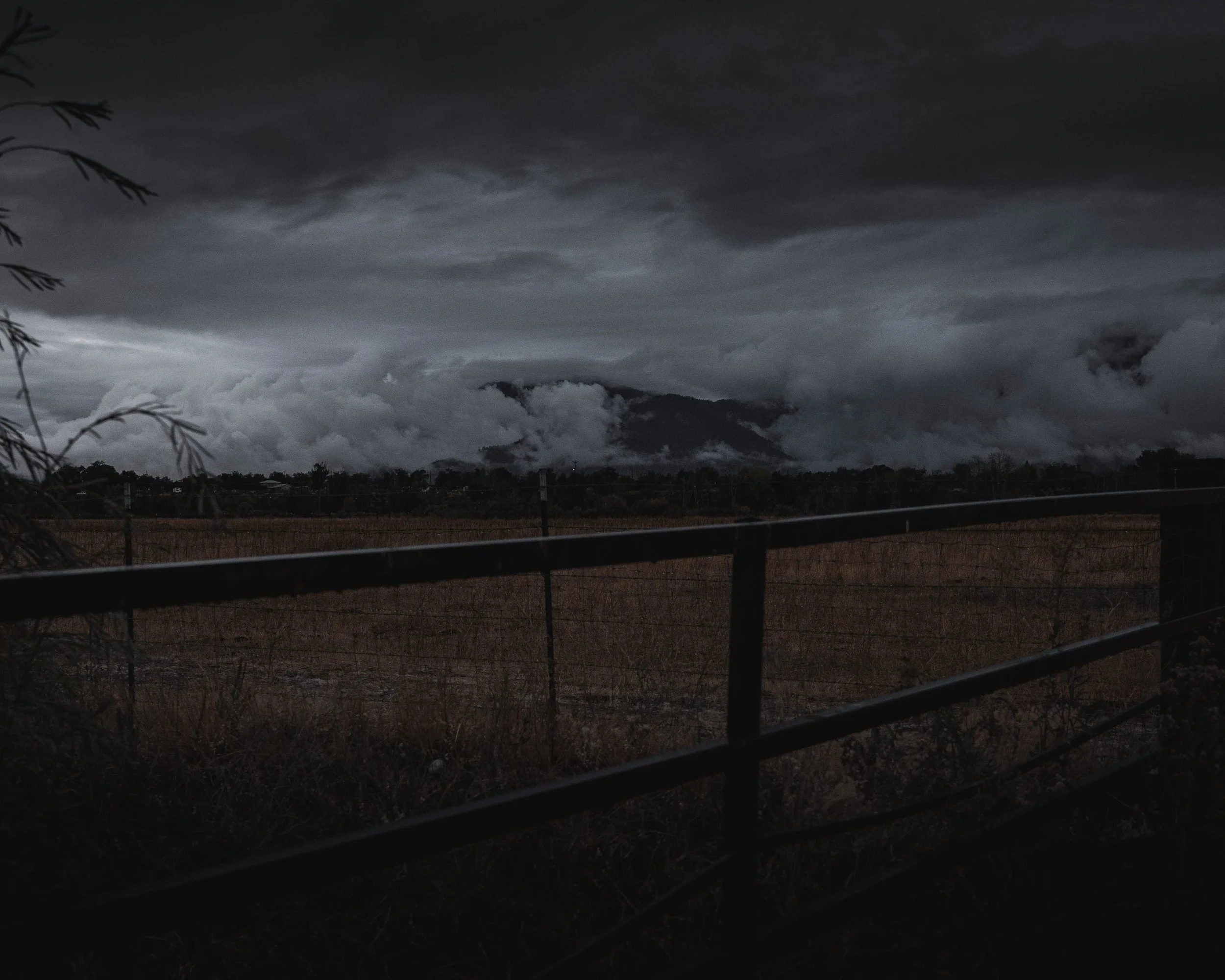 Dark, cloudy sky over mountains with low clouds, viewed from behind a metal railing.