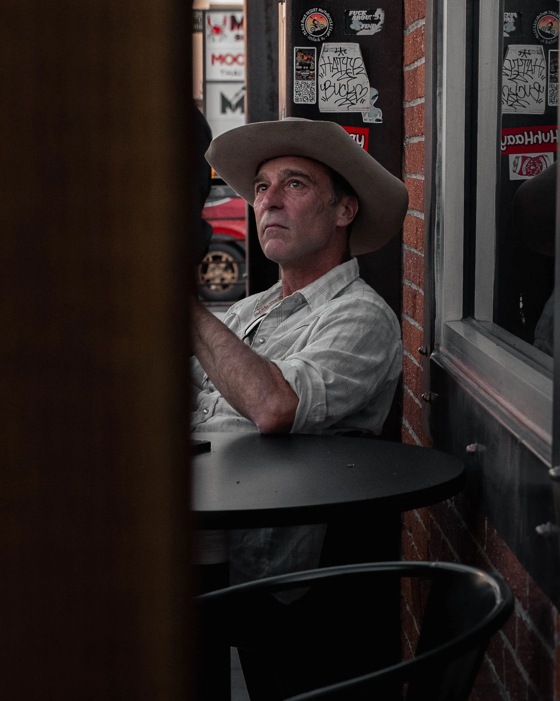A man wearing a western hat and a plaid shirt is sitting near a brick wall with stickers, next to a window. Part of a dark object is visible in the left foreground, and a black surface is in the lower part of the image.