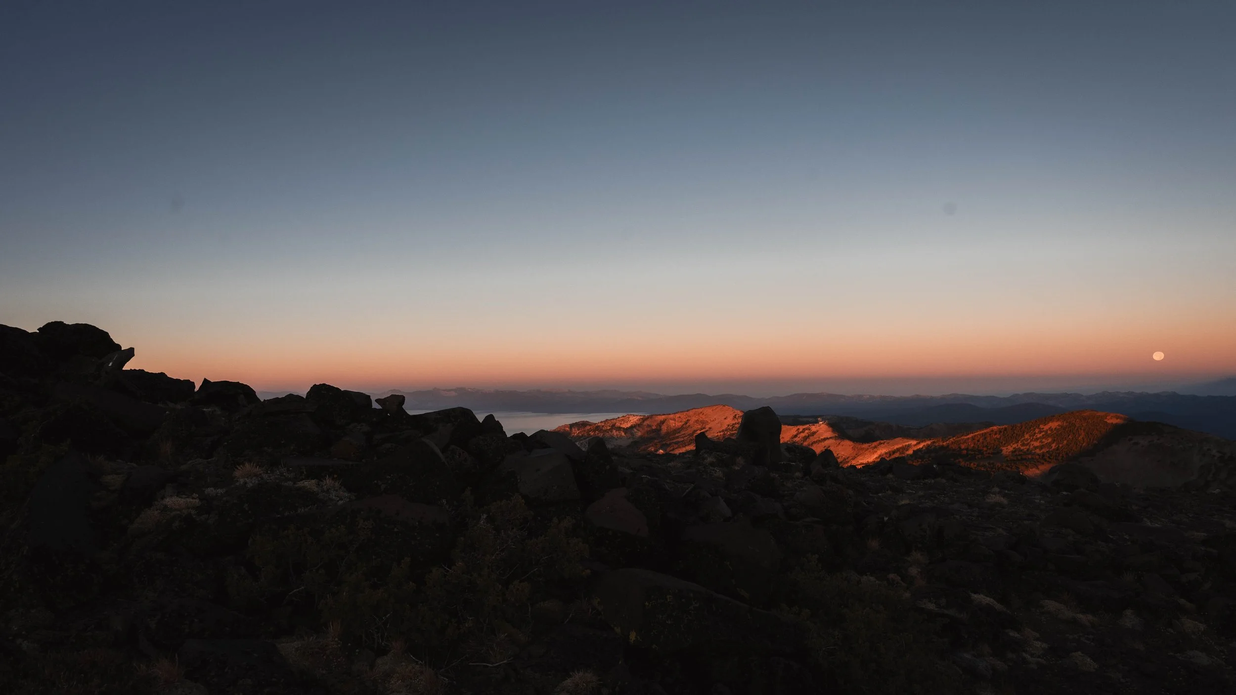 Sunset over rocky mountain landscape with a full moon in the sky.