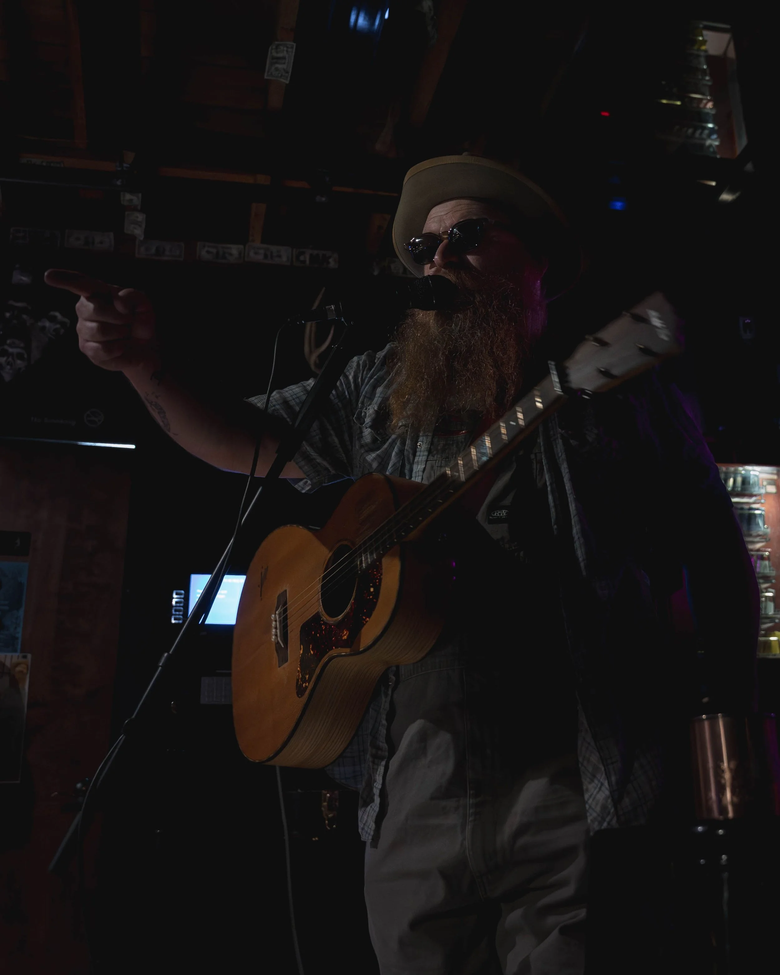 A bearded man wearing sunglasses, a beige hat, and a plaid shirt playing an acoustic guitar and pointing with his right hand, in a dimly lit room with shelves of CDs and posters.
