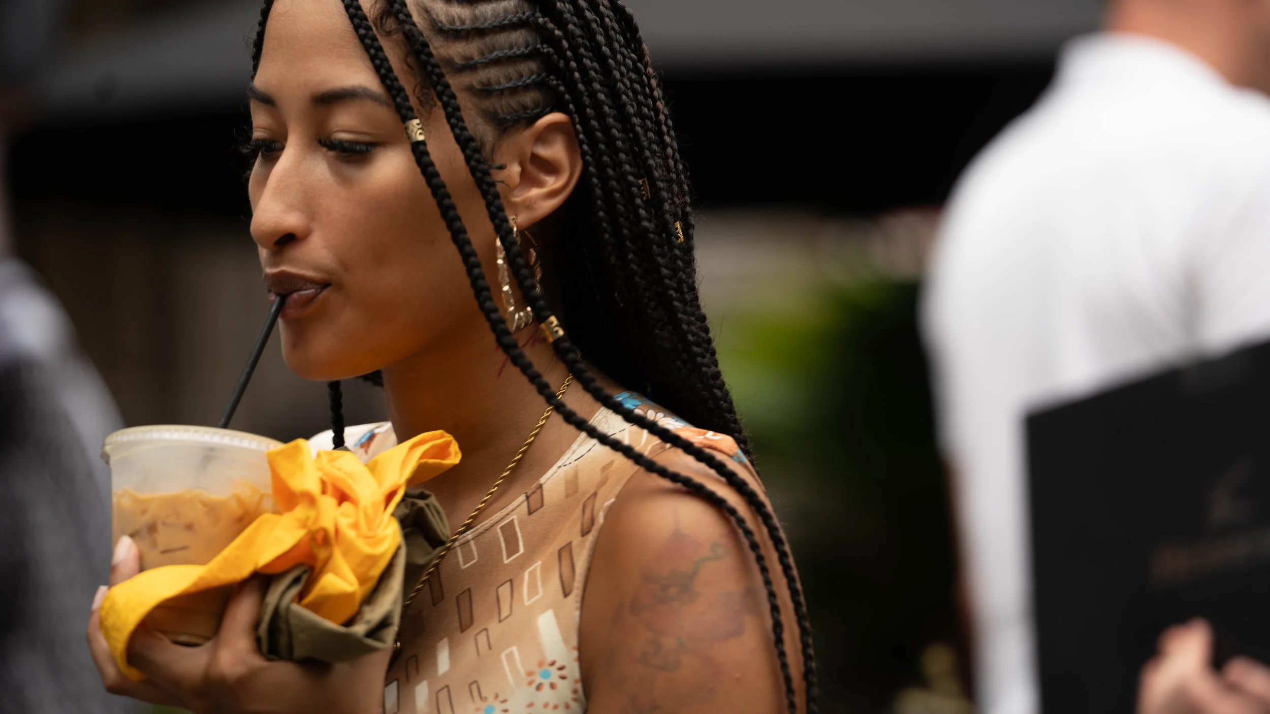 A woman with braided hair and earrings is drinking a beverage through a straw and holding a bouquet of yellow flowers.