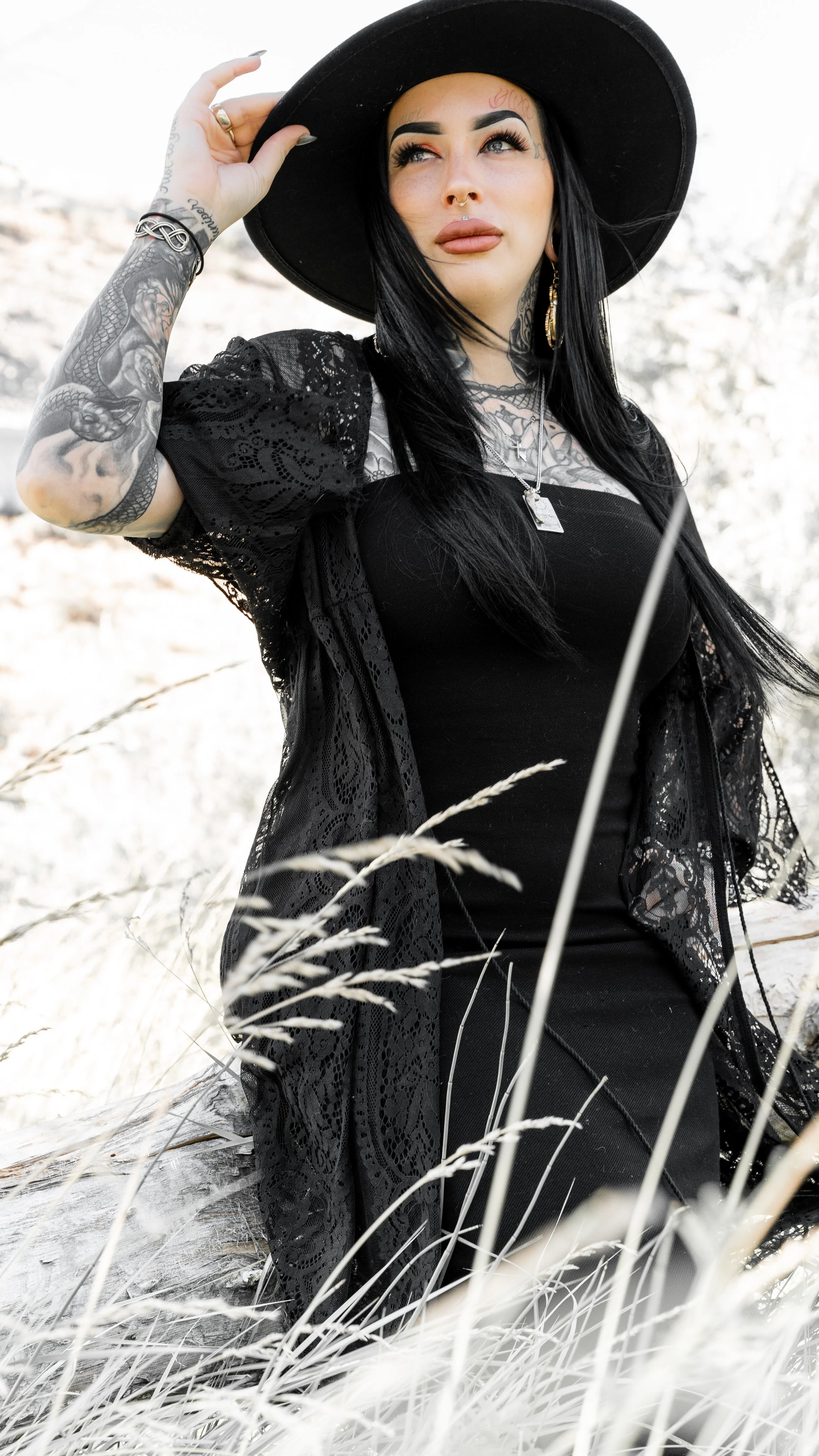 A woman with long dark hair, tattoos, and piercings wearing a black dress and wide-brimmed hat, sitting outdoors in a field of dry grass.