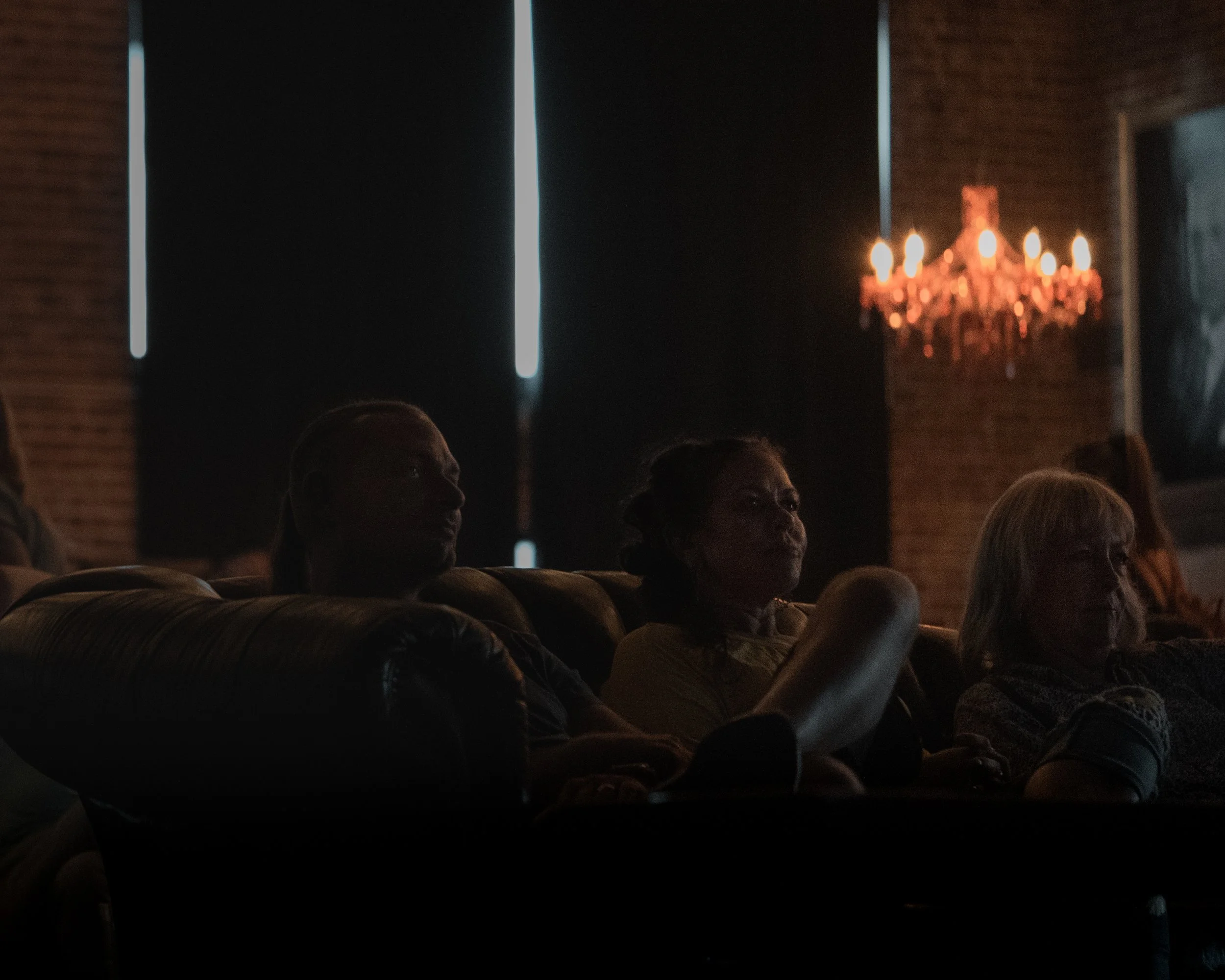 A dark room with a chandelier and people sitting in the audience, looking towards the stage or screen.