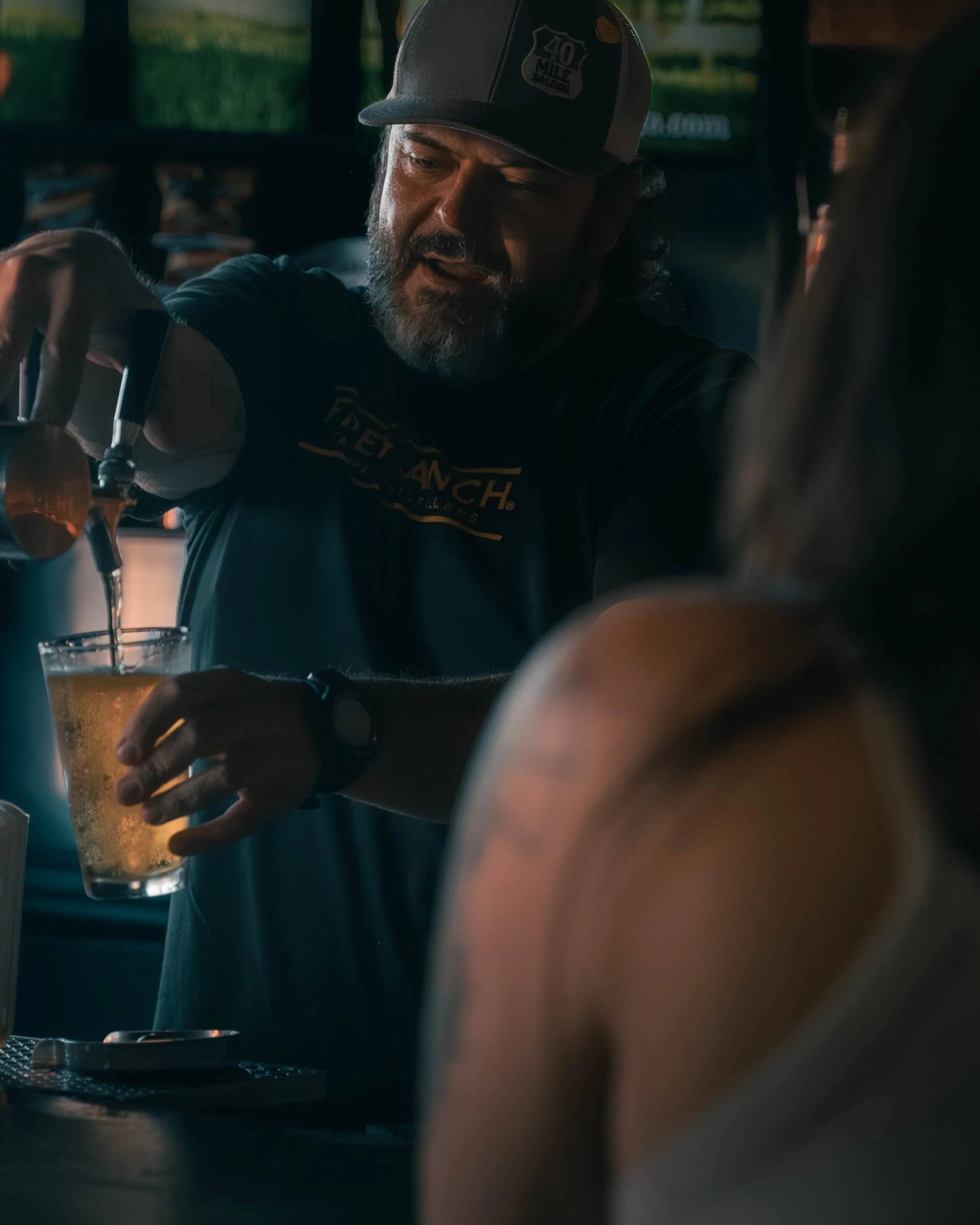 A man with a beard wearing a baseball cap pouring beer from a tap into a glass in a bar or pub setting.