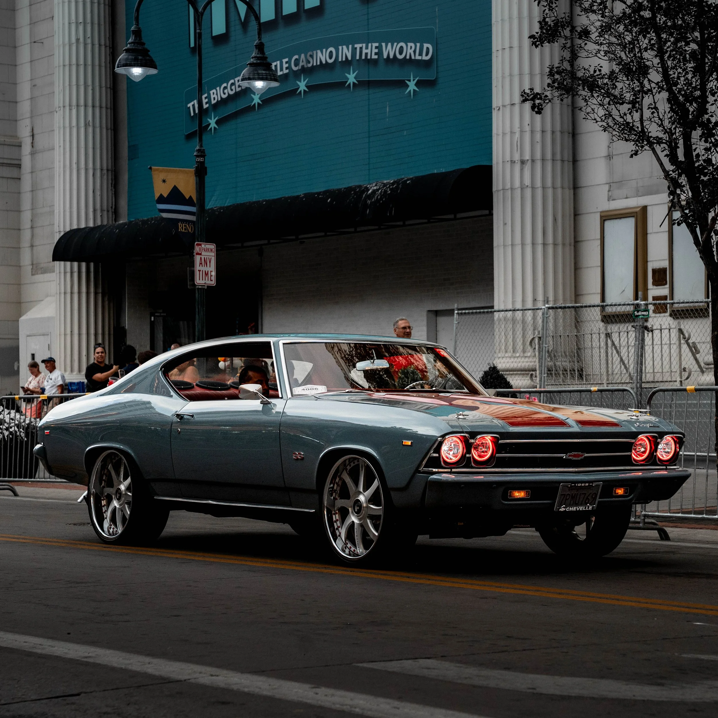 A classic muscle car, possibly a Chevrolet Corvette, parked on a city street in front of a building. The car has a sleek, gray body with red interior visible through the windows, and badge details at the rear. The background features a large blue bil