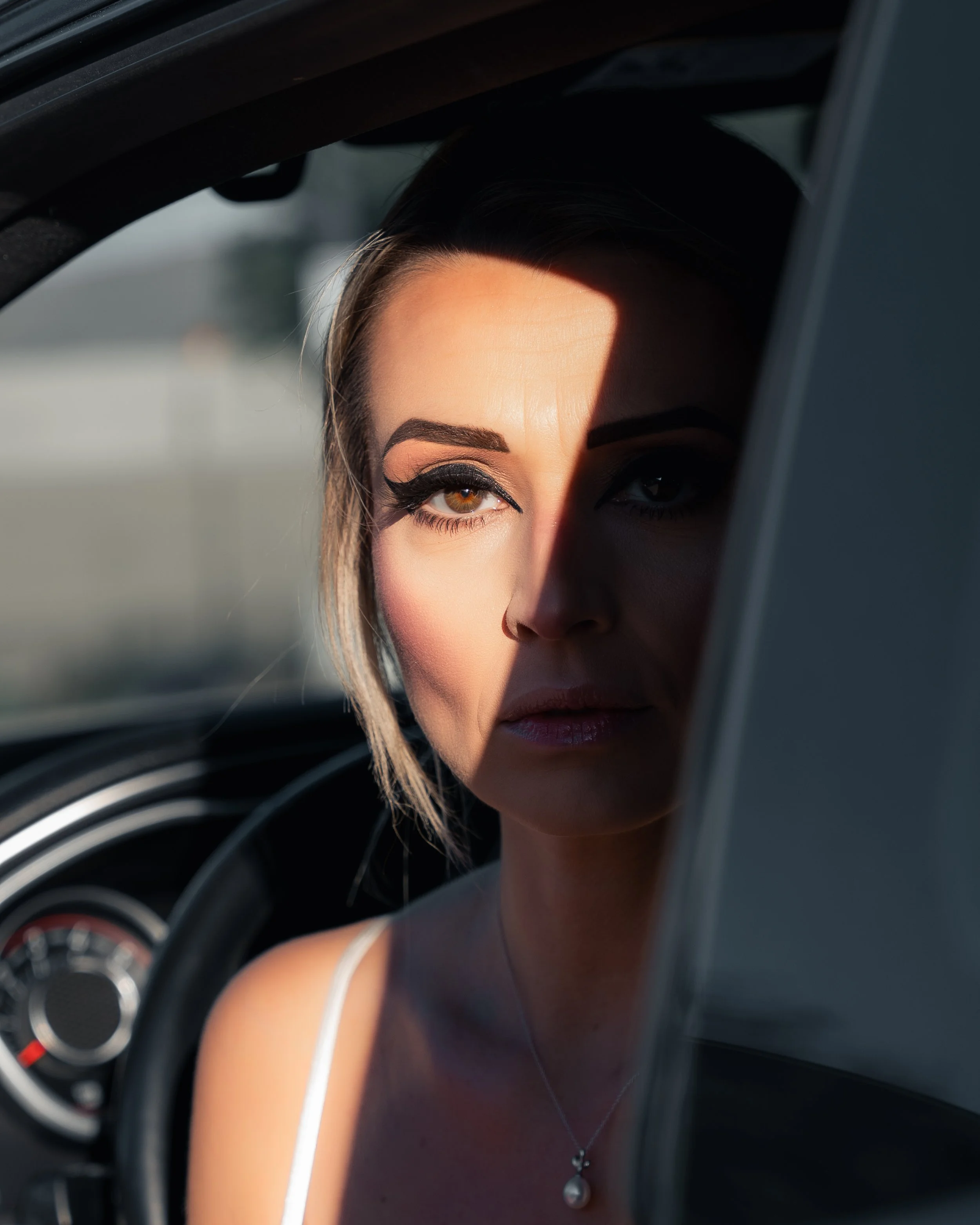 Close-up of a woman's face and upper body with makeup, seen through the open window of a car, with part of the dashboard and steering wheel visible.