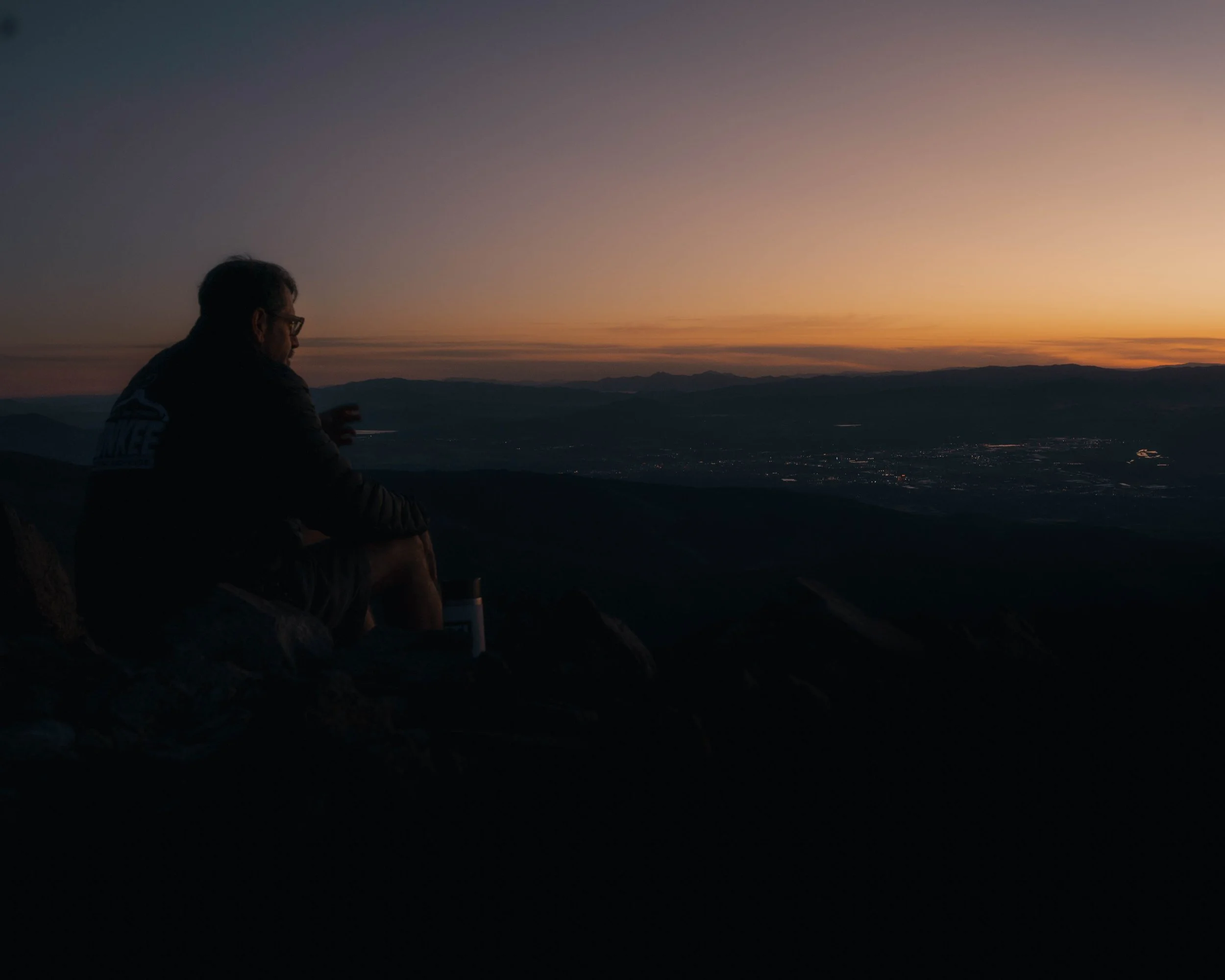 A person sitting on a mountain or hilltop during sunset, looking at the distant horizon with mountains and a valley below.