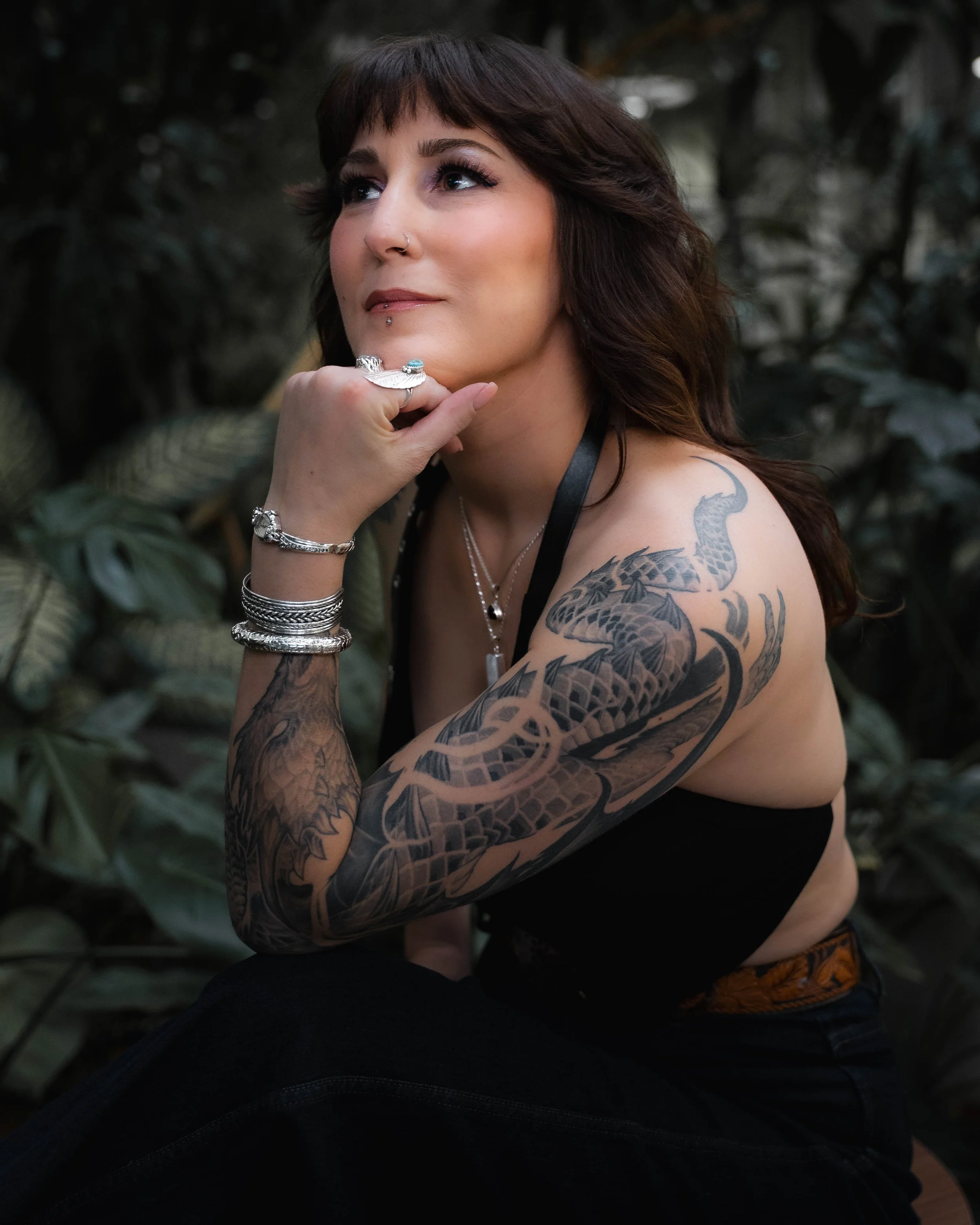 A woman with tattoos on her arm, wearing rings, and jewelry, resting her chin on her hand, in a natural outdoor setting.
