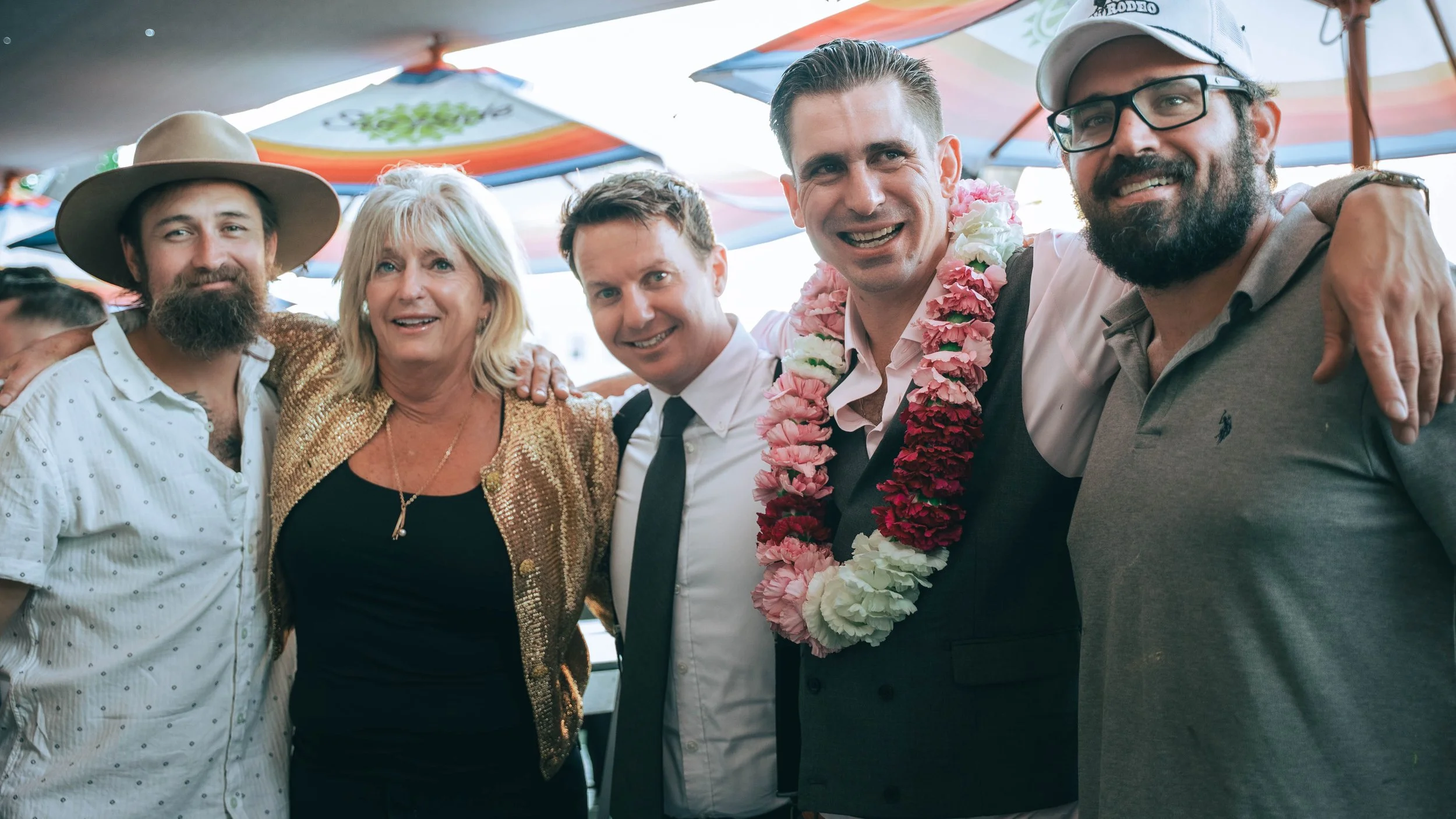 Group of six smiling people at an outdoor gathering, under colorful umbrellas, with one person wearing a flower lei and another wearing a large hat, all with their arms around each other.