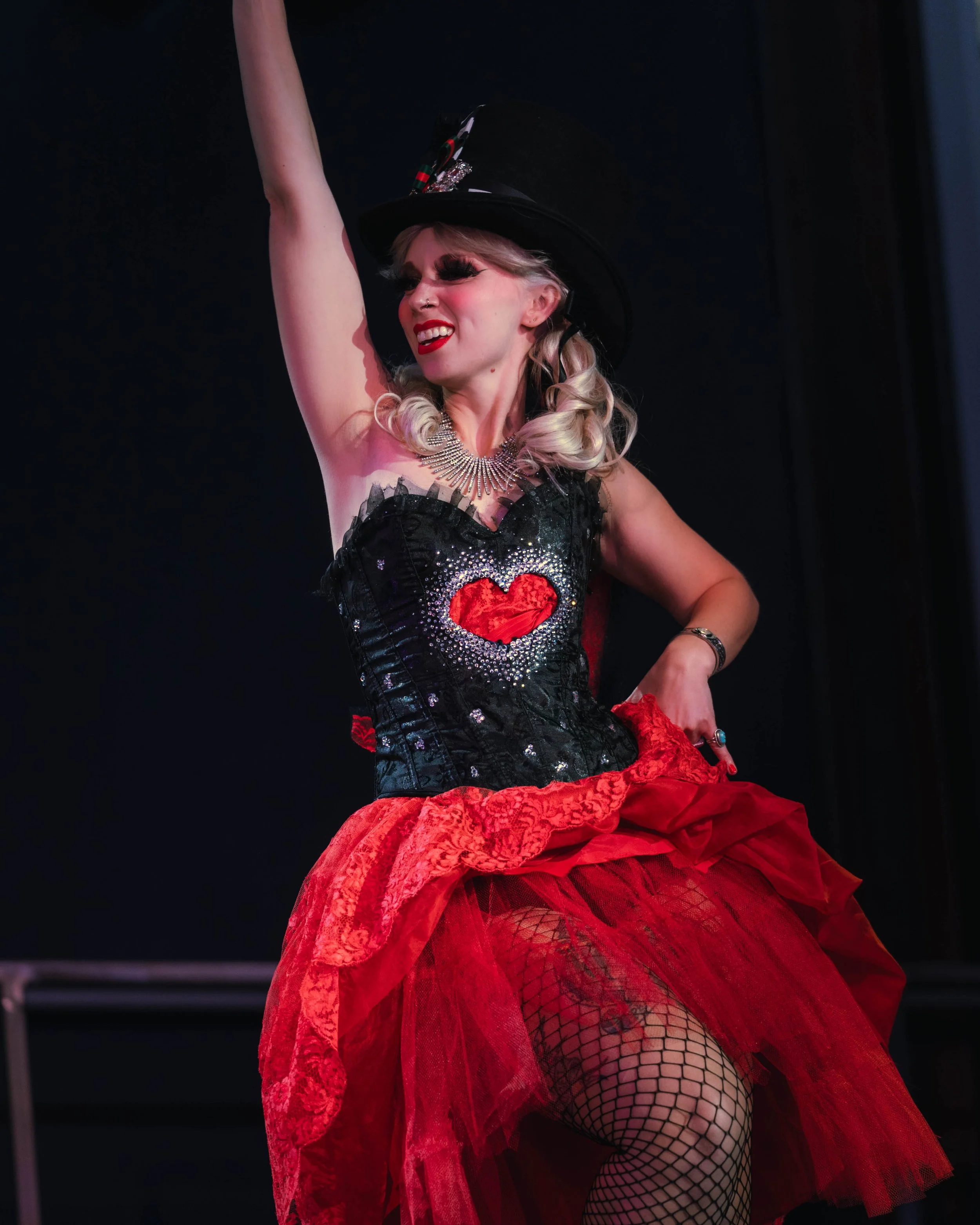A performer with blonde hair styled in loose curls, wearing dramatic makeup, a black glittery bodice with a red heart design, a large statement necklace, a tall black hat with embellishments, and fishnet stockings, dancing on stage with one arm raise