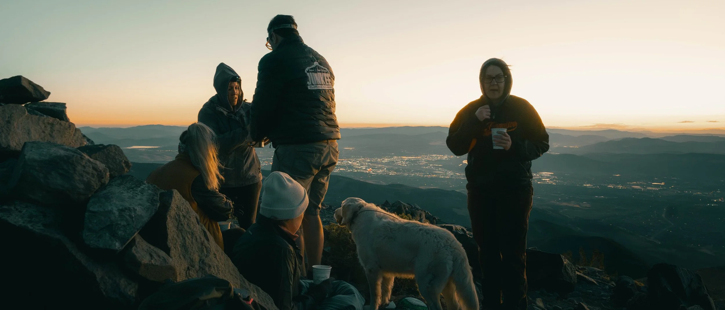 Group of people on a mountain summit during sunset, with some sitting and others standing, eating and drinking, with a dog, rocks in the foreground, and a panoramic view of a valley below.