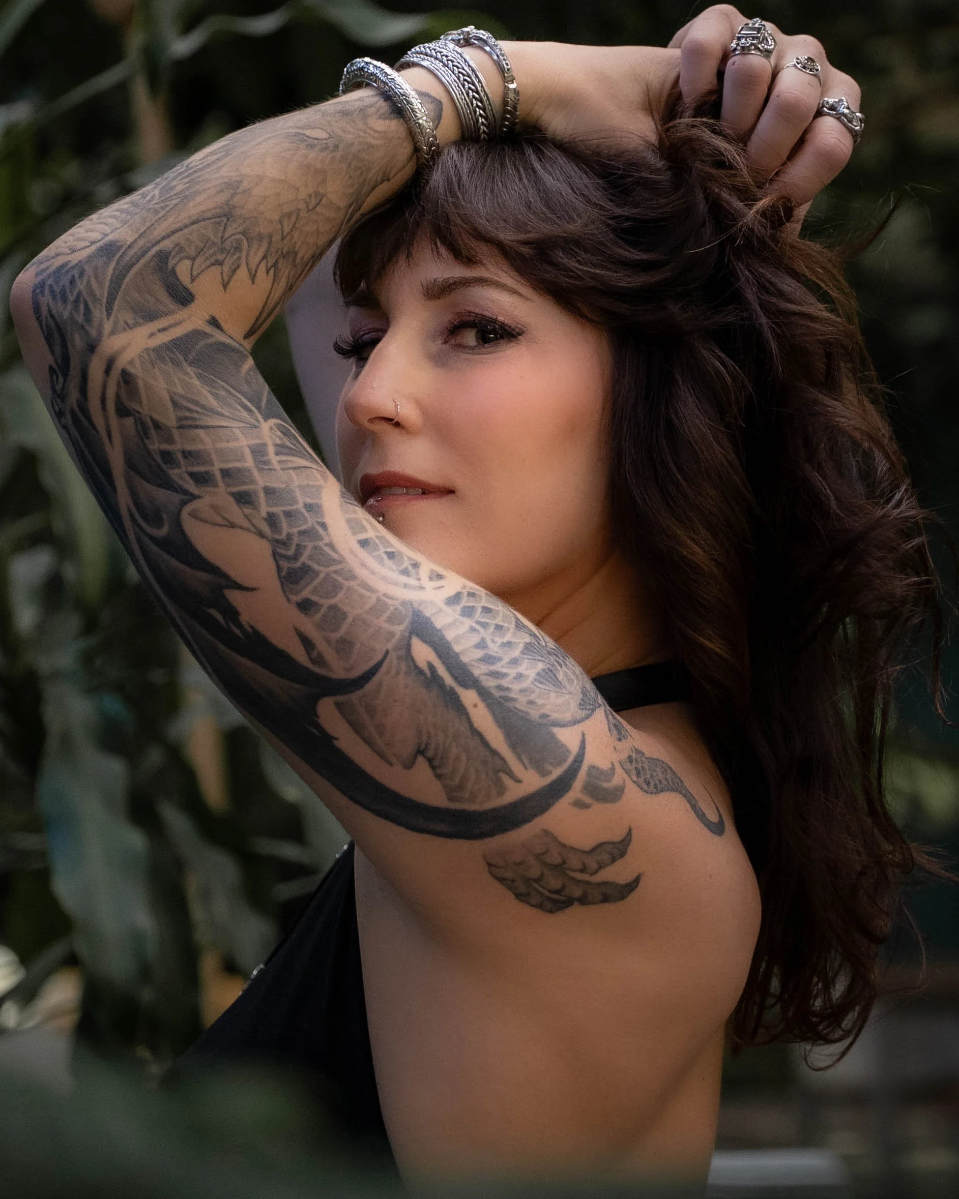 A woman with dark brown hair and light skin, wearing multiple rings and bracelets, poses with her left arm raised, showcasing her tattooed arm. She has a nose piercing and is partially smiling, with lush greenery in the background.