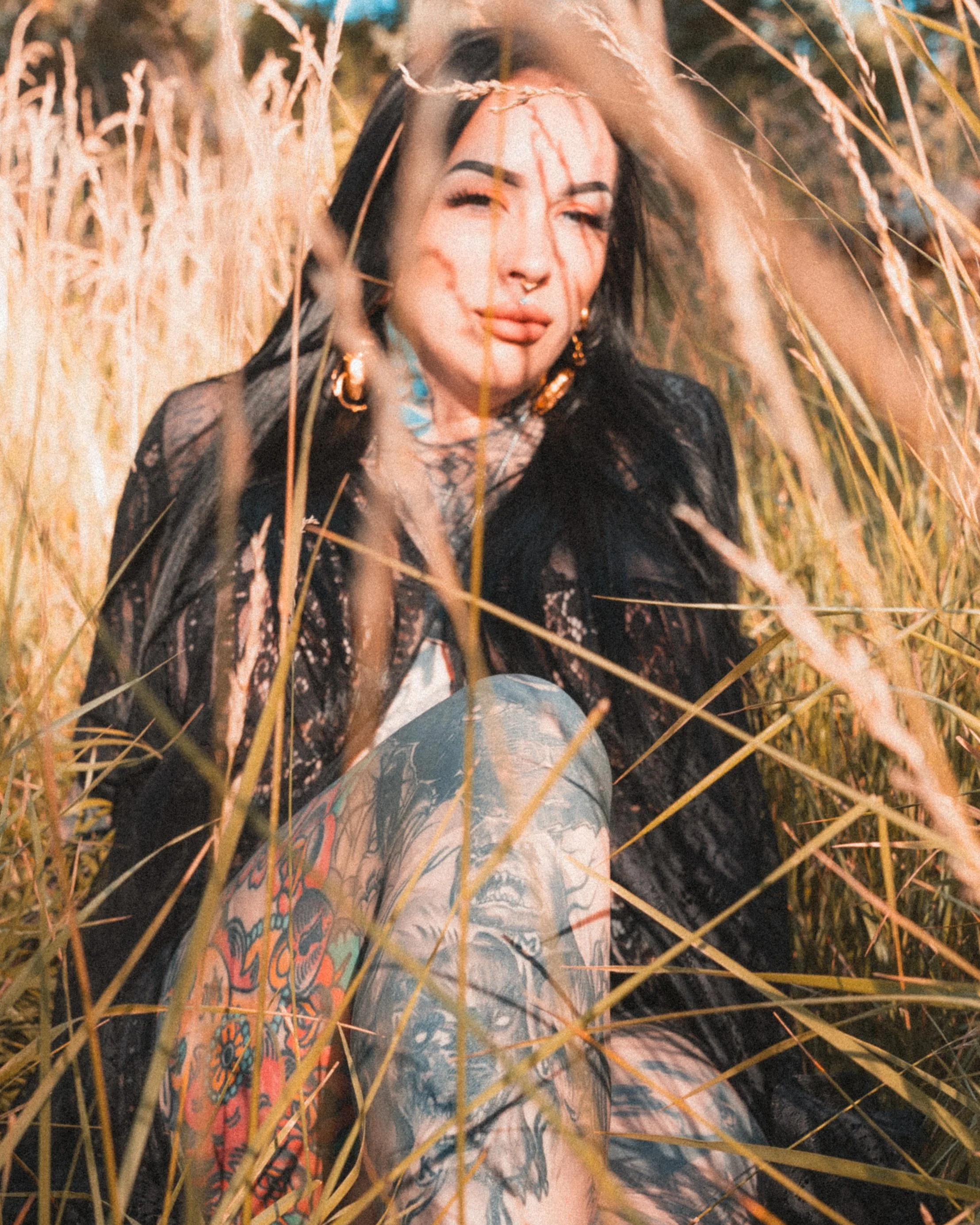 A woman with black hair and tattoos on her arms and chest sitting among tall, golden grass.