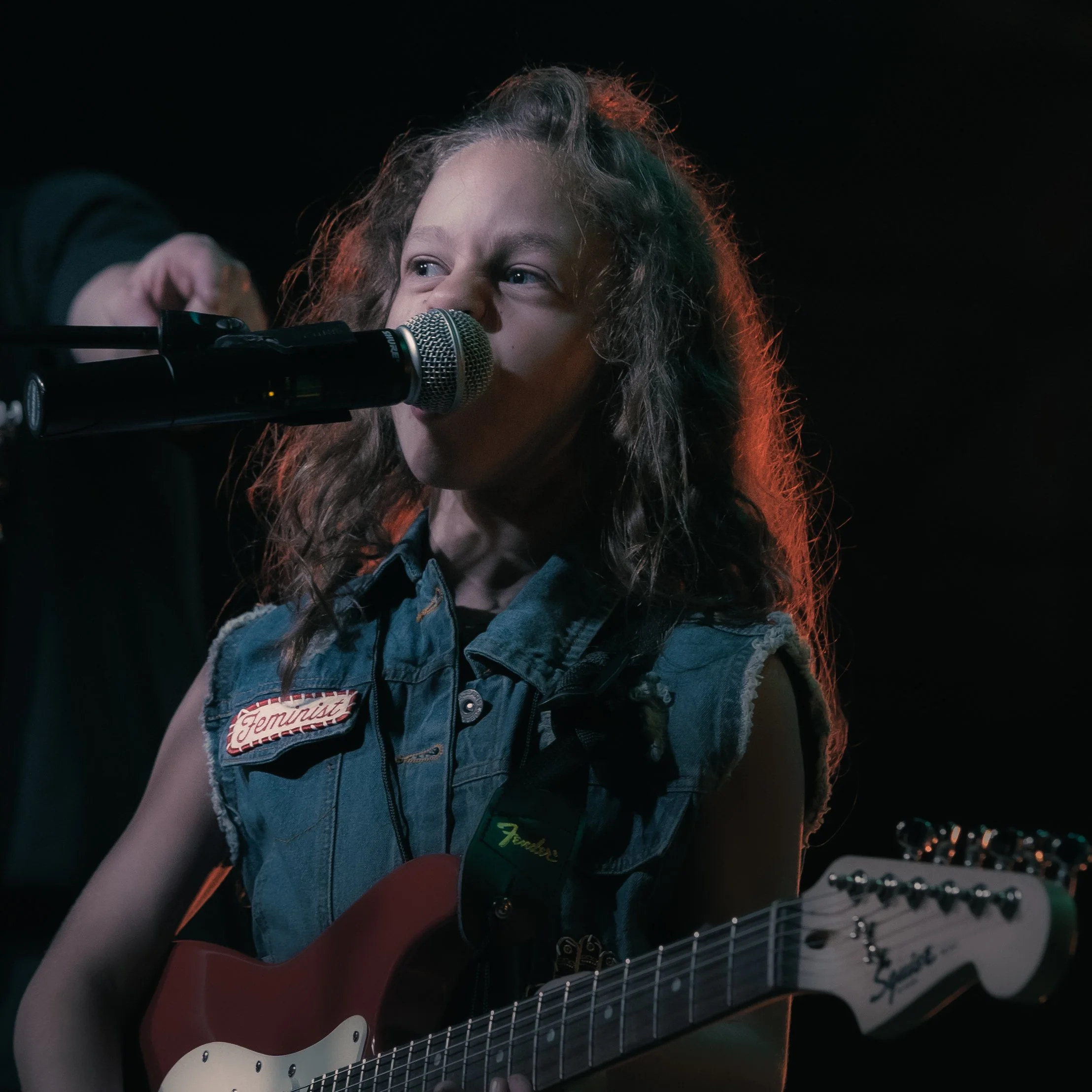 Close-up of a person singing into a microphone while playing an electric guitar, wearing a denim vest with patches in a dark setting.