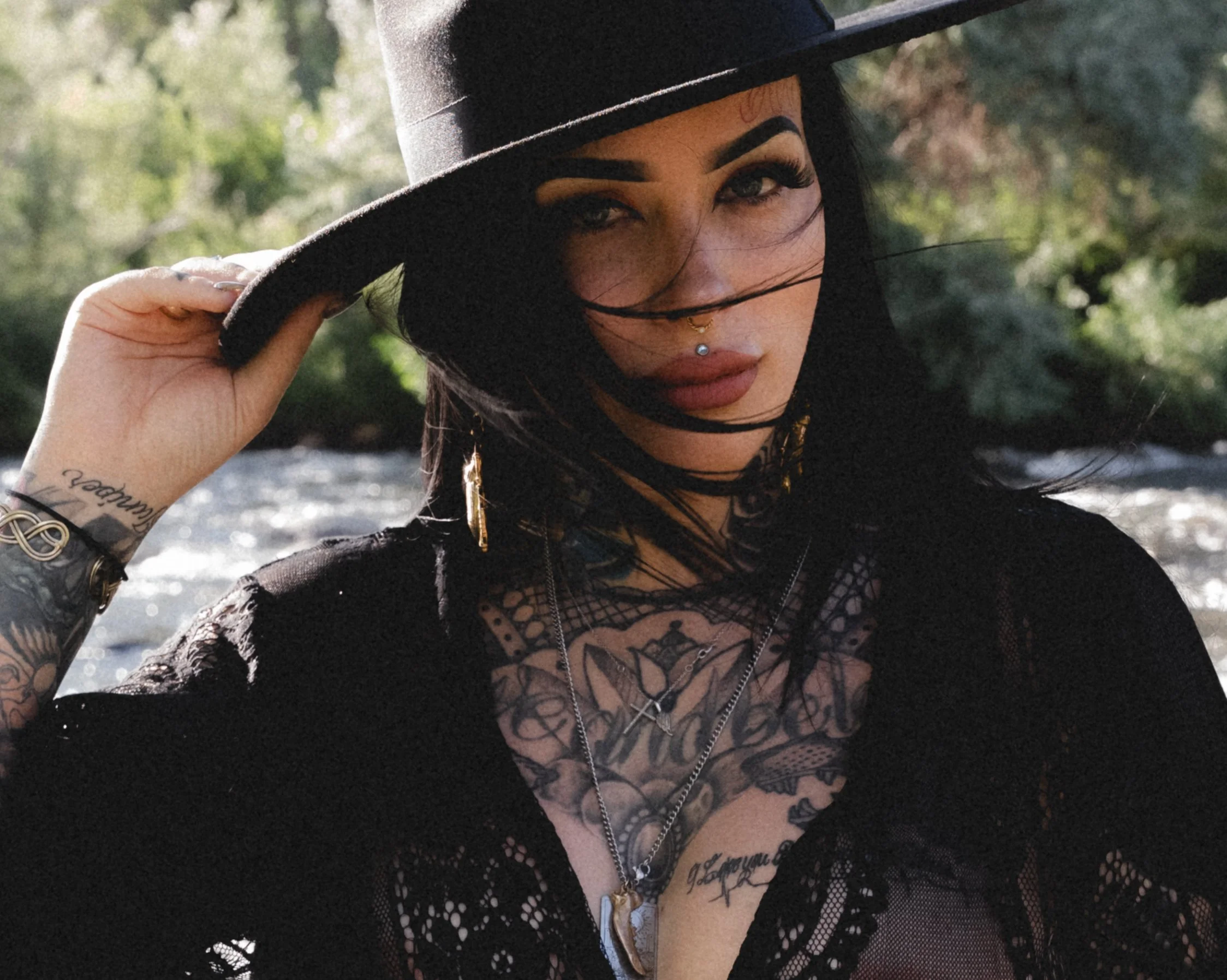 A woman with dark hair, dark makeup, and tattoos, wearing a black lace top, a black hat, and layered jewelry, outdoors during daytime.
