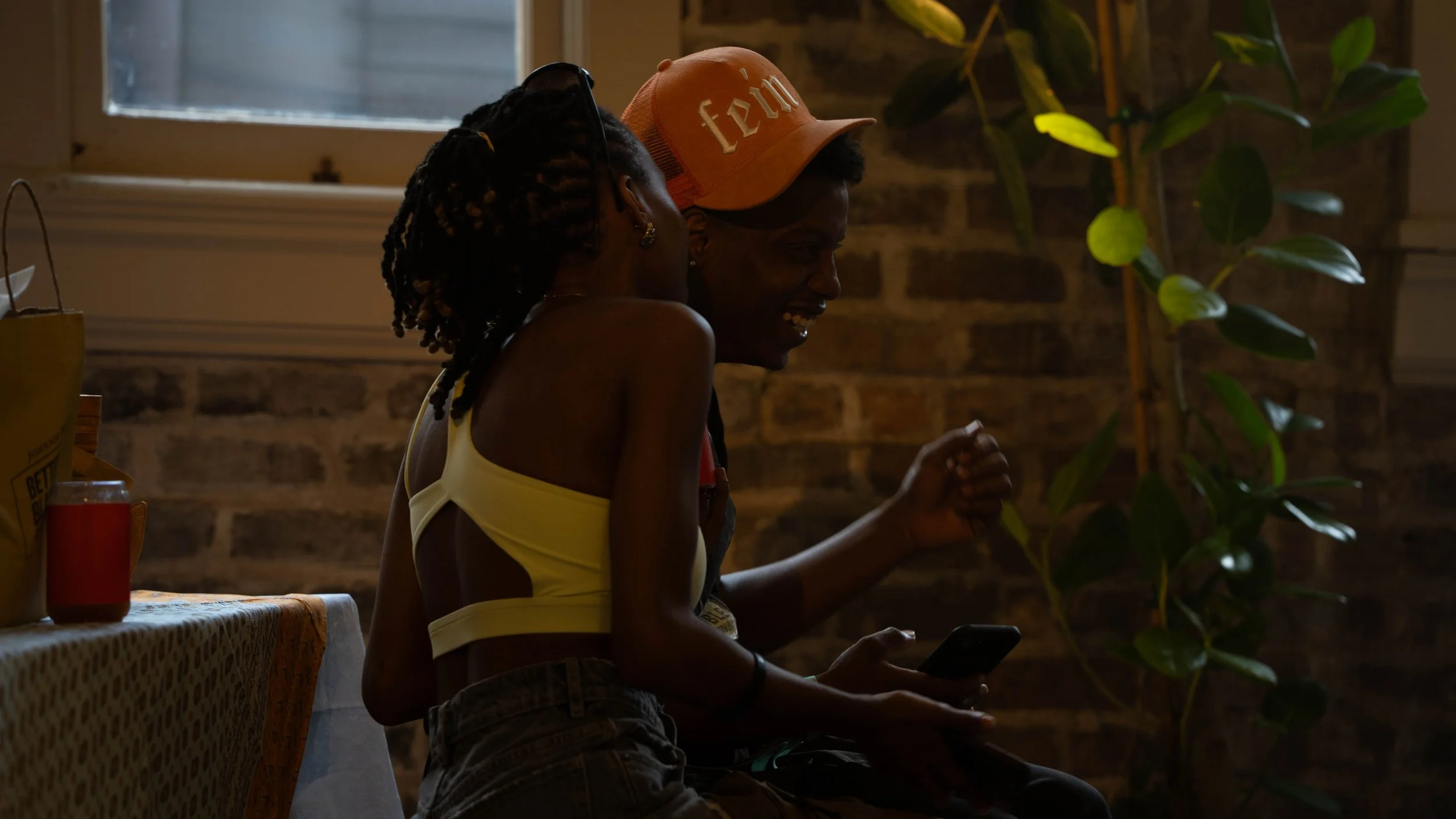 Two young women sitting together indoors, one wearing a yellow top and a cap, the other with dreadlocks and earrings, both smiling and looking at a smartphone. There are plants and a brick wall in the background.