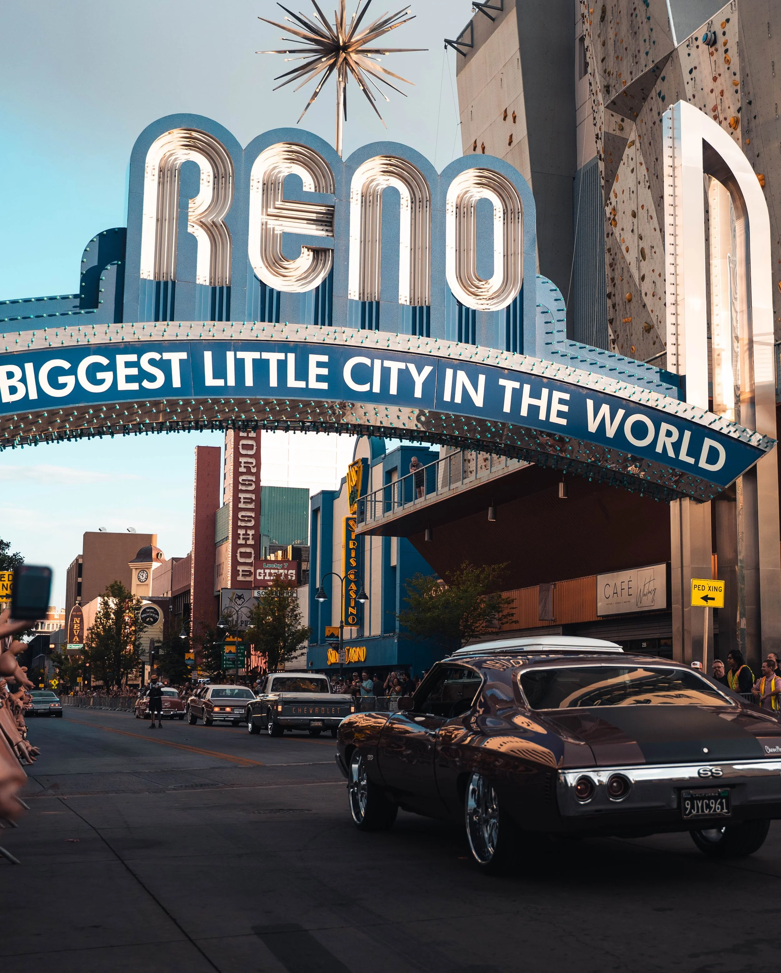 Vintage cars driving under a Neon Sign that reads "Welcome" with the subtitle "Biggest Little City in the World."