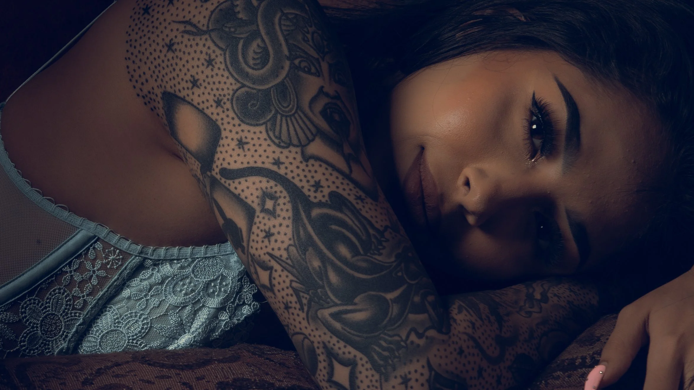 A woman with dark hair and eye makeup, lying down with her eyes closed. She has a large tattoo on her right shoulder and arm featuring various designs and dots. She is wearing a lace or crochet top.