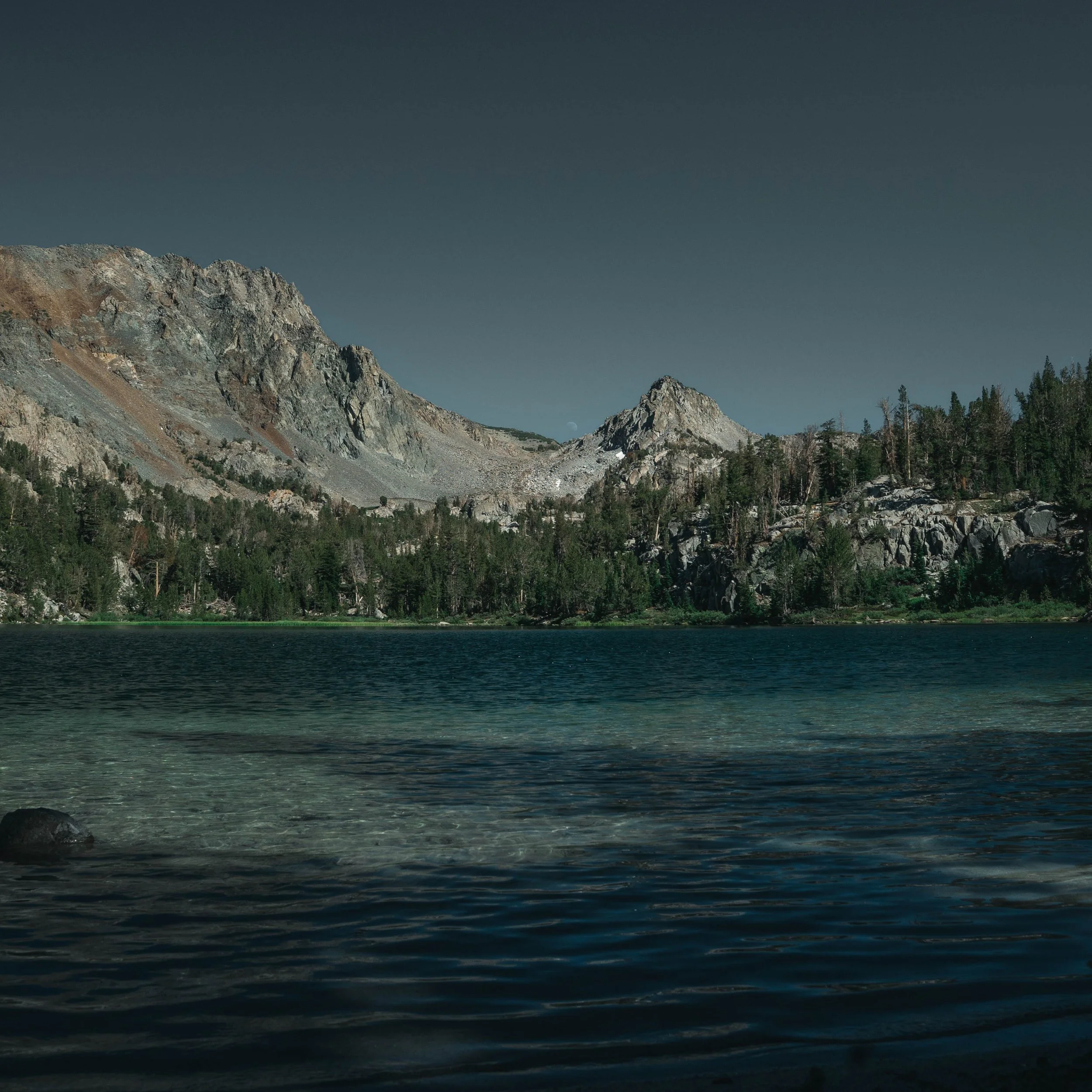 Scenic view of a mountain range with rocky peaks, evergreen trees, and a lake in the foreground under a dark sky.